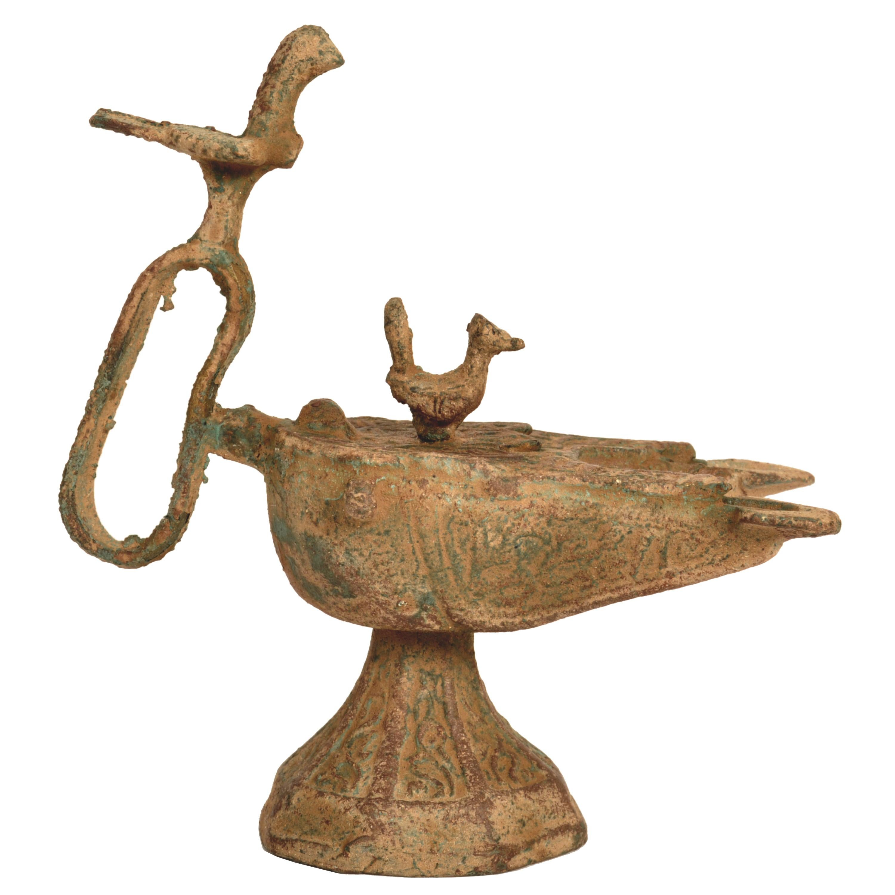 A 12th Century Islamic bronze oil lamp, Eastern Persia, Khurasan.
The cast bronze body of compressed globular form, raised on a trumpet shaped foot, the hinged cover with a sculpted bird finial and  there is a corresponding finial to the loop