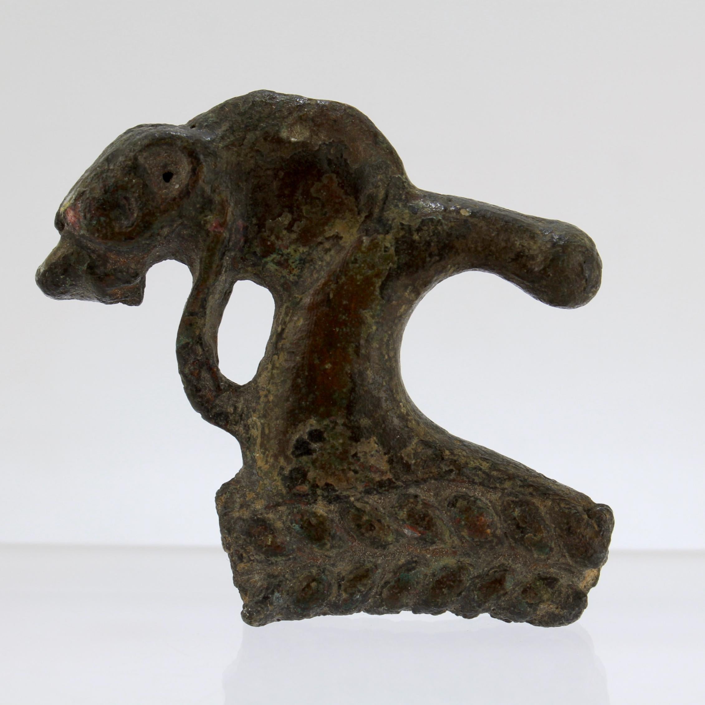 An antique or ancient Roman bronze element. 

This piece was an element of a vase or possibly handle to something larger. 

It has the head of a wolf or lion mounted on a curved handle supported by a section of braided ring. 

With a thick