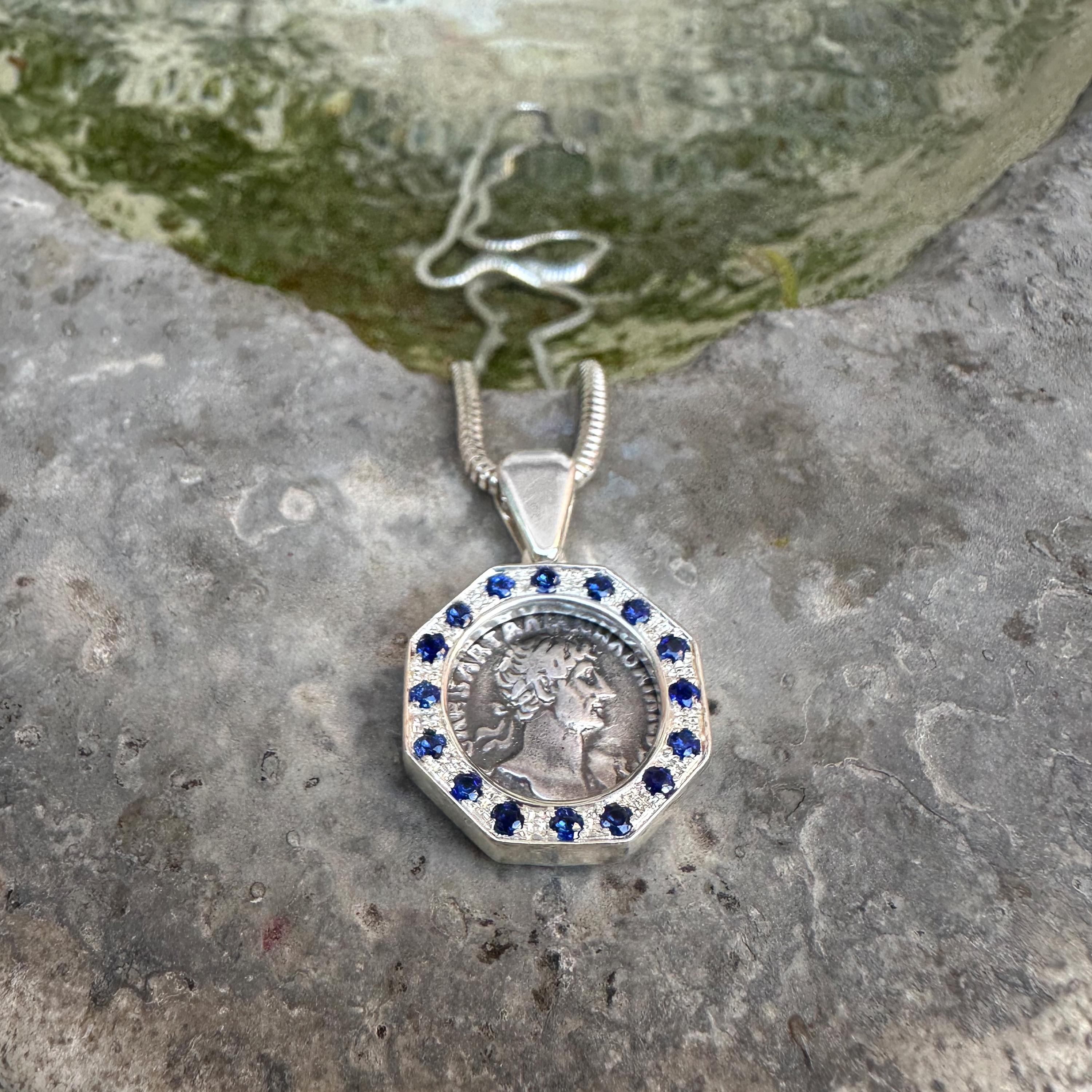 This sterling silver pendant holds an authentic Roman coin dating back to the 2nd century AD, depicting Emperor Hadrian, surrounded by 16 Sapphires. The reverse side showcases the goddess Aeternitas who, in the religion of Ancient Rome, was the