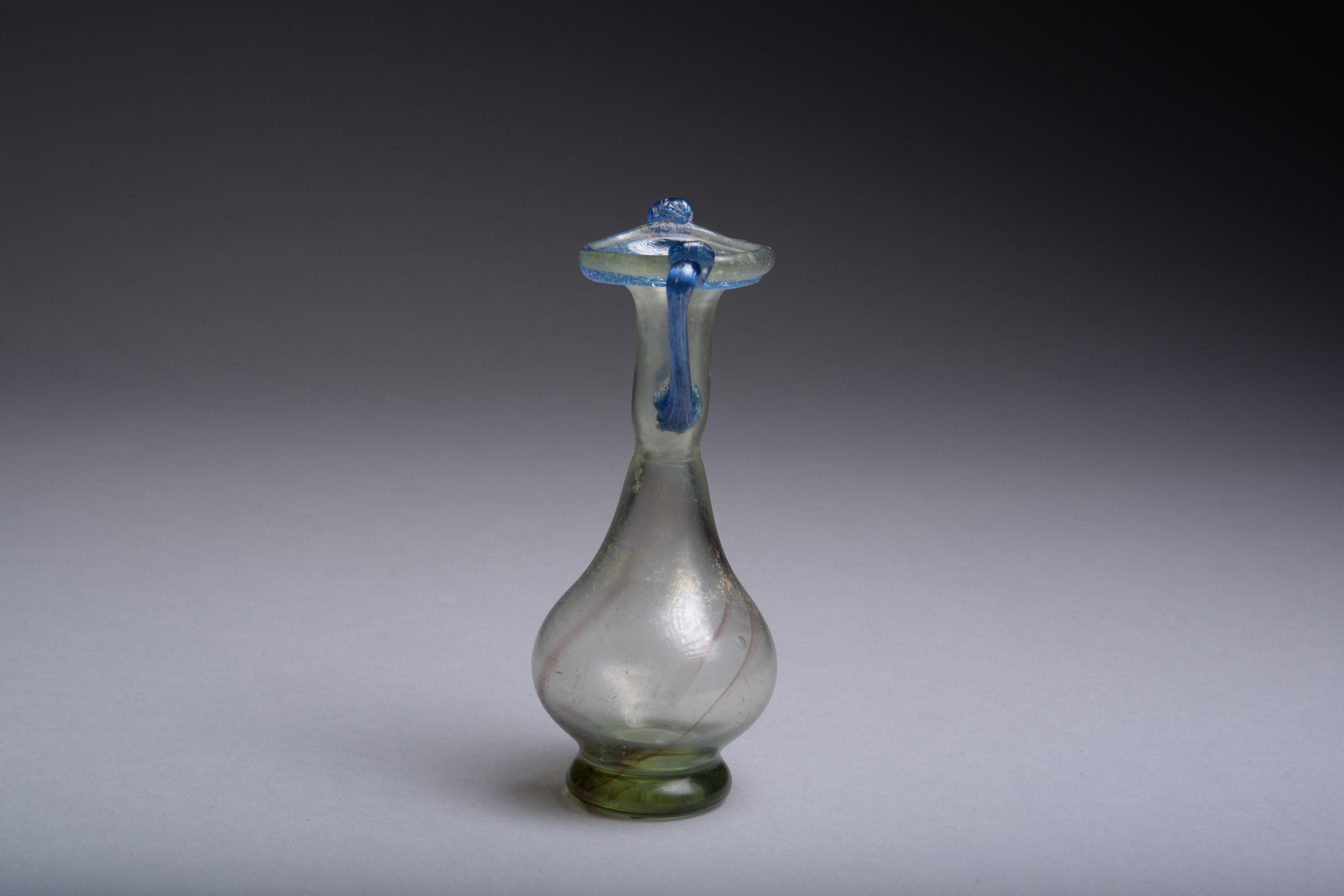 A Roman glass bottle, dating to the 3rd-4th century AD.

Blown from pale green glass with streaks of purple. Of elegant form, with thick circular base, rounded body, long neck and two applied blue glass handles, a trail running under the