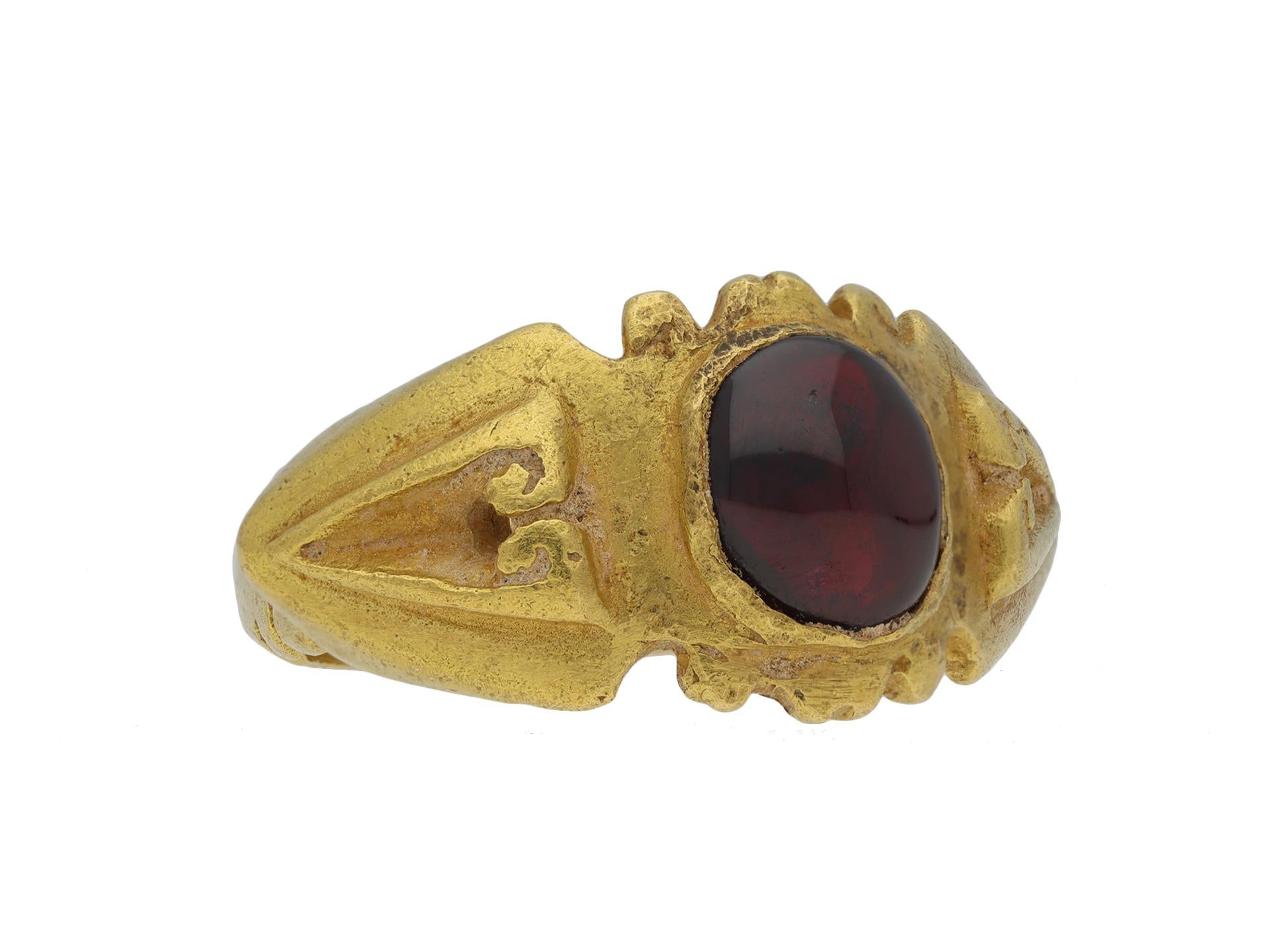 Ancient Roman Syrian garnet signet ring. Set with one central oval cabochon garnet in a rubover setting, to an embellished signet design featuring a shaped bezel, geometric motifs, leading to raised angular shoulders with a scrolling pattern and