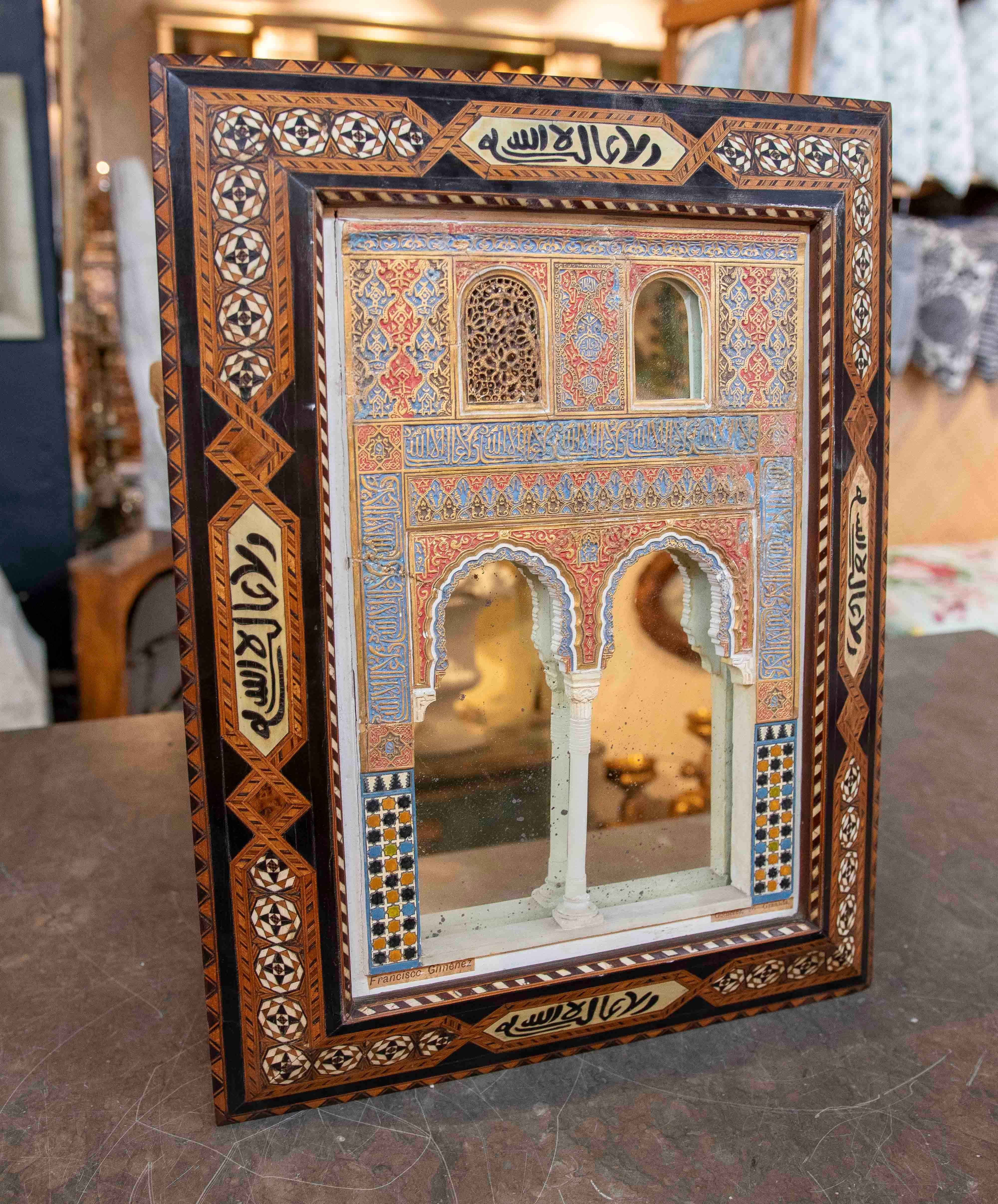 Exquise maquette artisanale de l'Alhambra, inspirée de l'architecture andalouse, avec un cadre en bois décoré de marqueteries géométriques, d'inscriptions arabes et de reliefs polychromes. Elle représente des arcs lobés et des tracés minutieusement