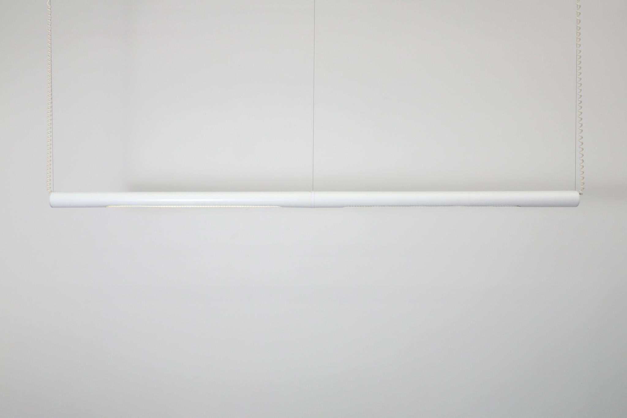 Lampada da lavoro fluorescente a doppio tubo, lunga e lineare, smaltata di bianco, sospesa da tre cavi con doppia spirale che si collega a ciascuna estremità. Ciascuna sezione misura circa 50
