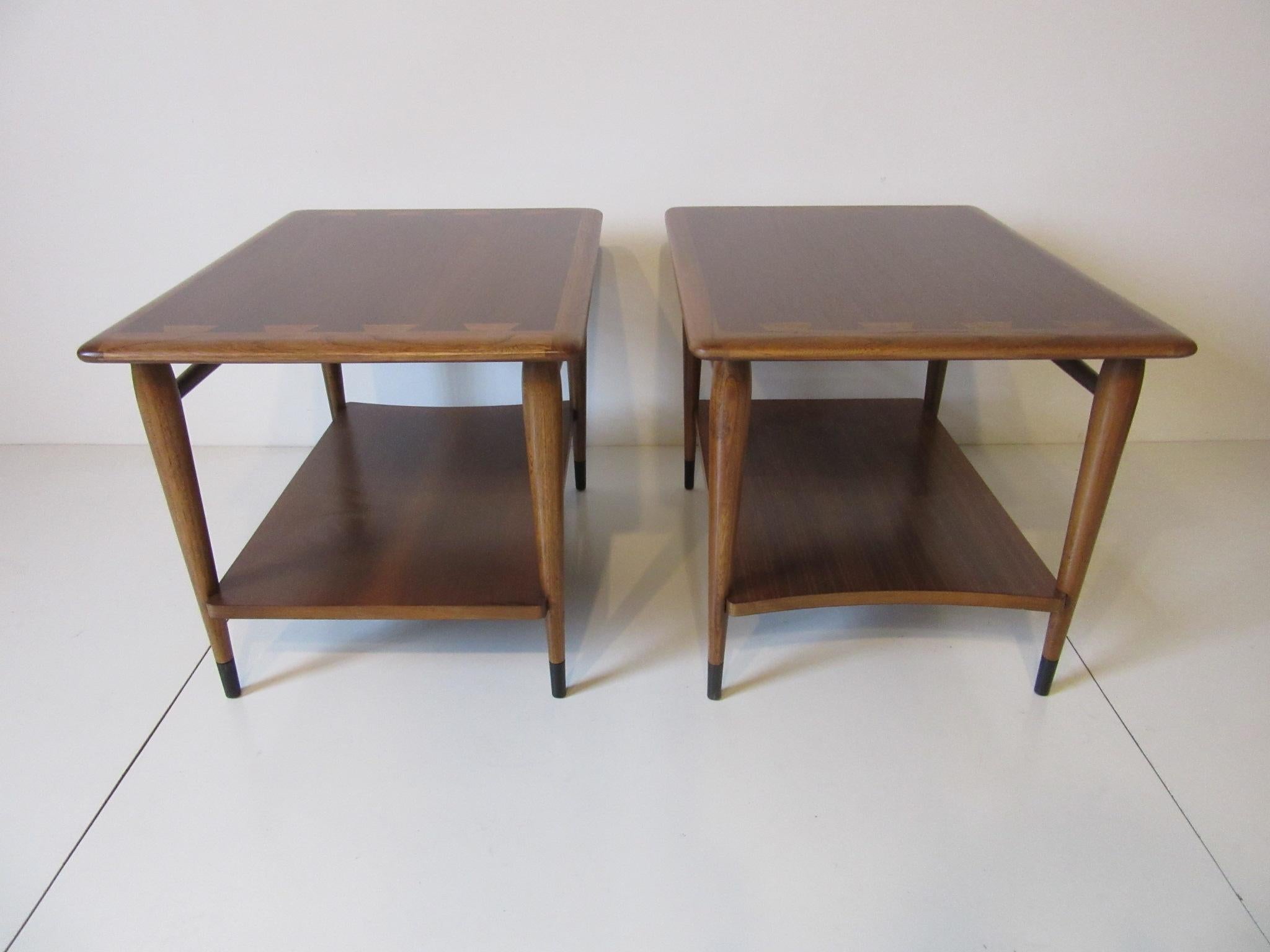 A pair of walnut end tables with lighter wood details to the edges, black lower leg caps and second shelve, one shelve is squared and the other has a curved edge . From the Acclaim collection and still retains the branded manufactures mark to the