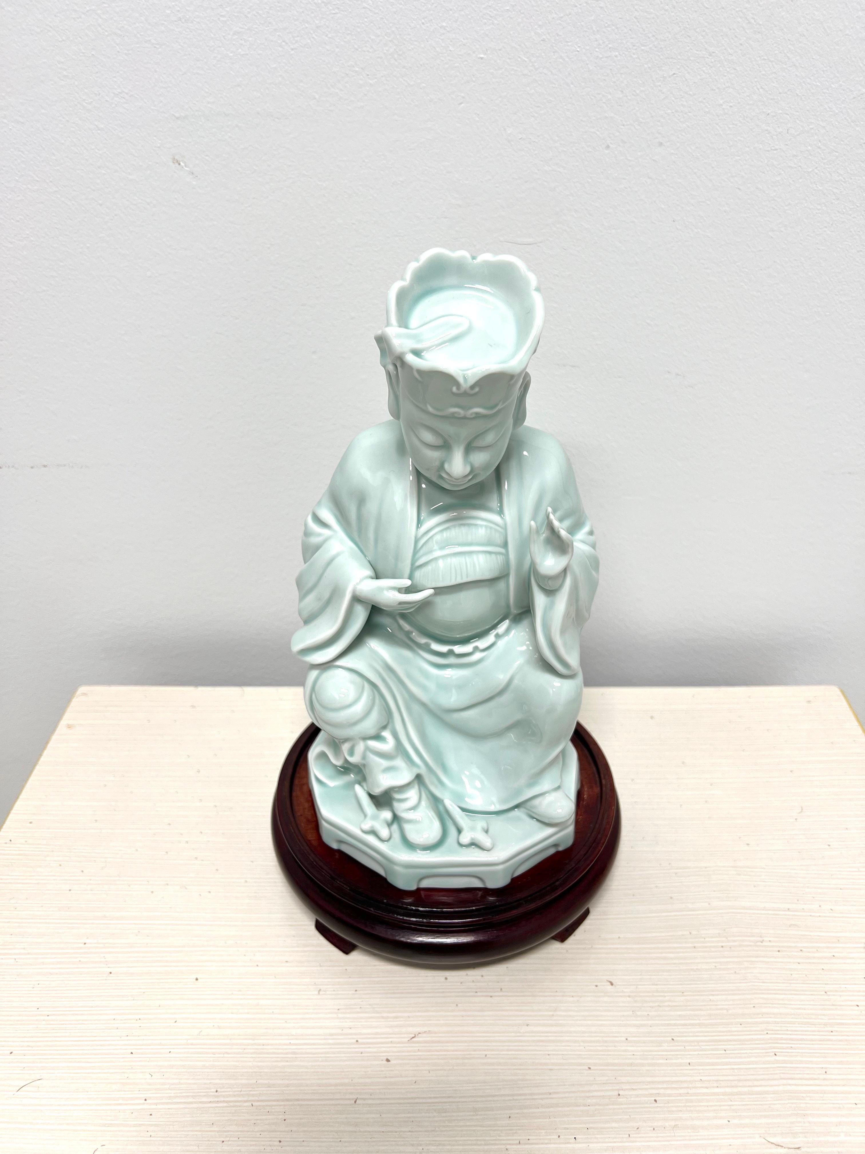 An Asian style Buddha figurine with wood stand by Andrea by Sadek. Hand painted, light blue color glazed porcelain of a seated Buddha, and a separate decoratively carved round wood base. Made in Japan, in the late 20th Century.

Measures:  Overall: