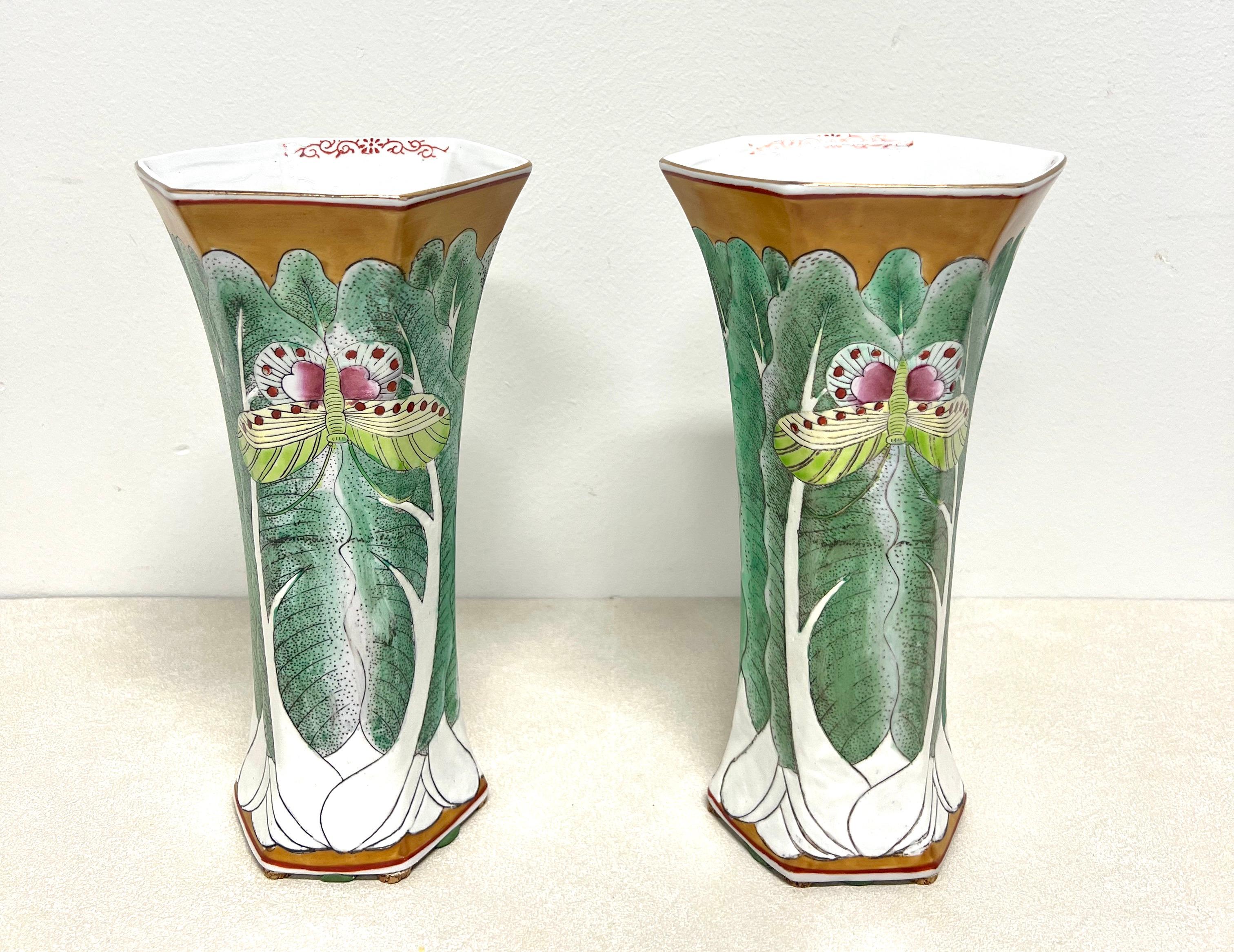 A pair of Asian Chinoiserie style table vases by Andrea by Sadek. Hand painted famille vert porcelain with vivid colors, primarily greens, depicting bok choy & butterflies, gold trim, and a hexagonal shape. Made in China, in the late 20th