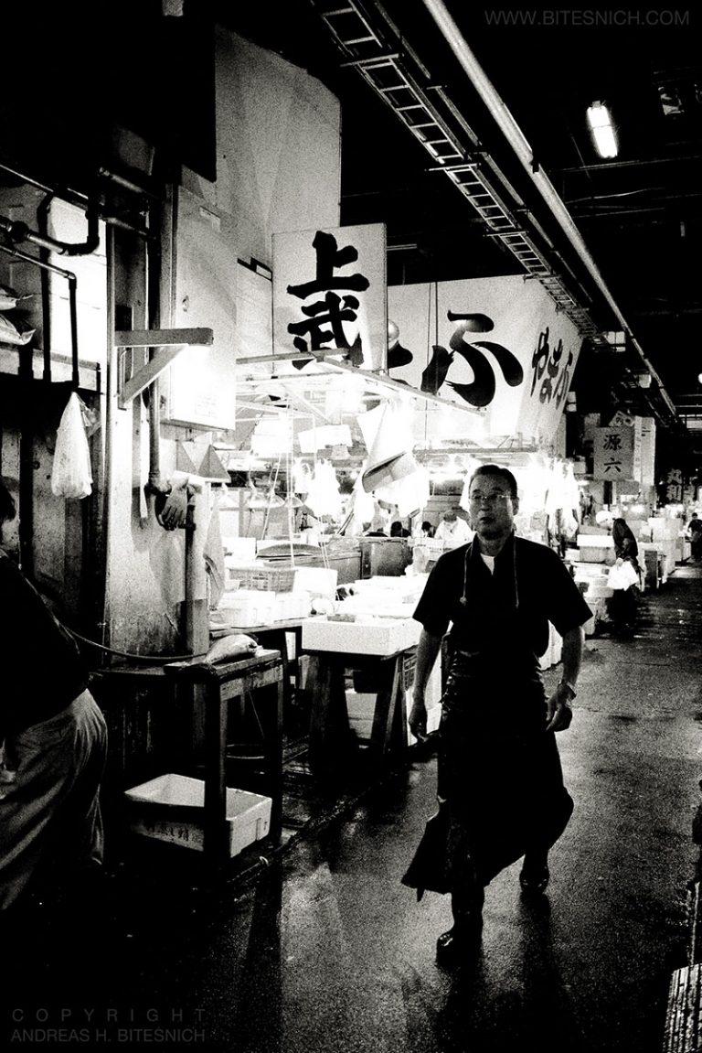 Andreas H. Bitesnich Black and White Photograph – Deeper Shades, Tokio – ein Mann, der auf einem Fischmarkt geht, mit Schildern im Hintergrund