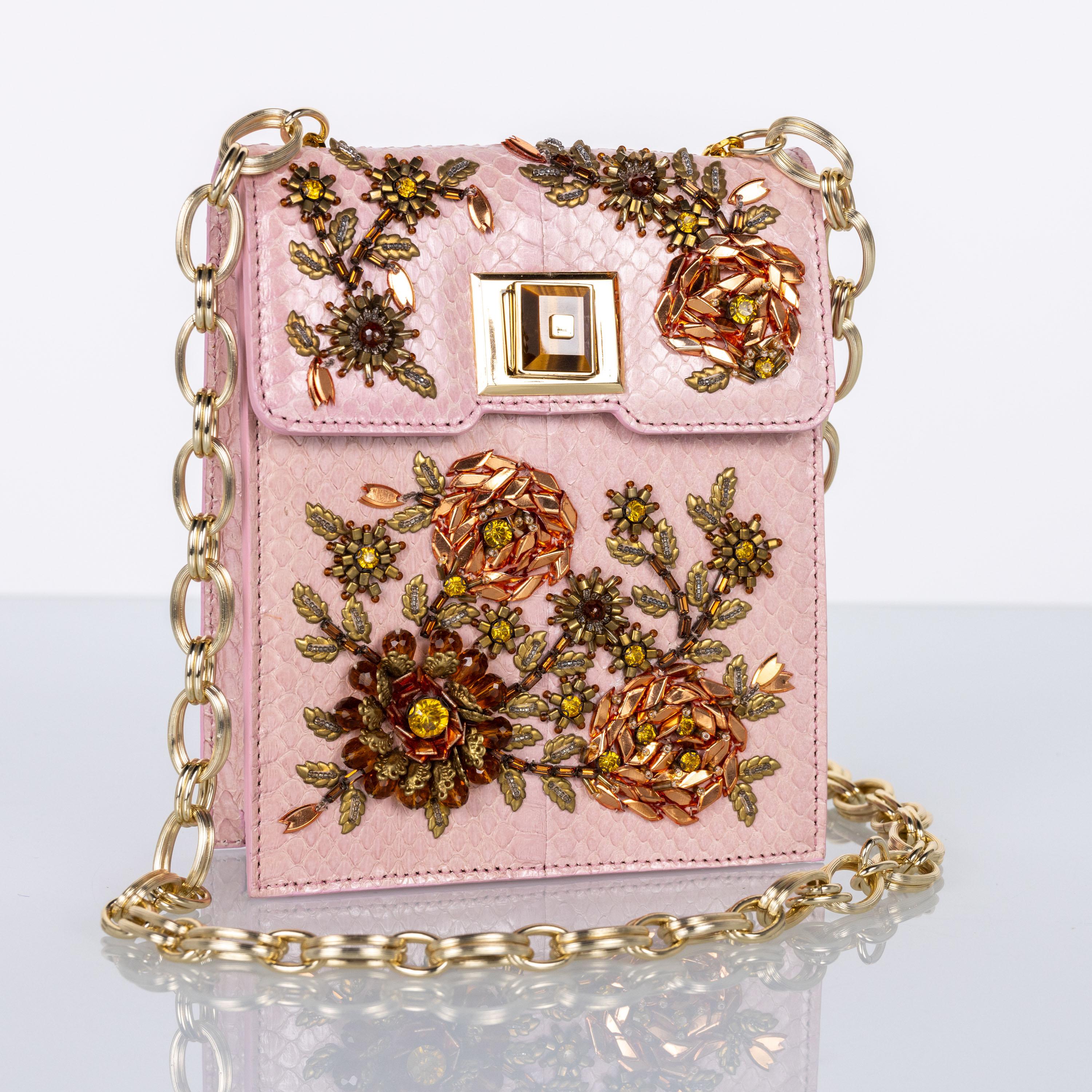 This exquisite Andrew Gn handbag features a pink snake leather base adorned with intricate floral embroidery, brought to life through beads, sequins, and metallic accents in gold and bronze tones. A long gold chain strap adds versatility, while the
