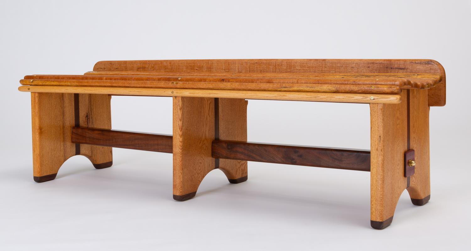 An American Studio Craft bench by Andrew Stauss showcases beautifully figured wood and contrasting grains. The slatted seat is gently curved with a low back panel, and each oak rail has rounded edges. The bench has three legs of live oak, bisected