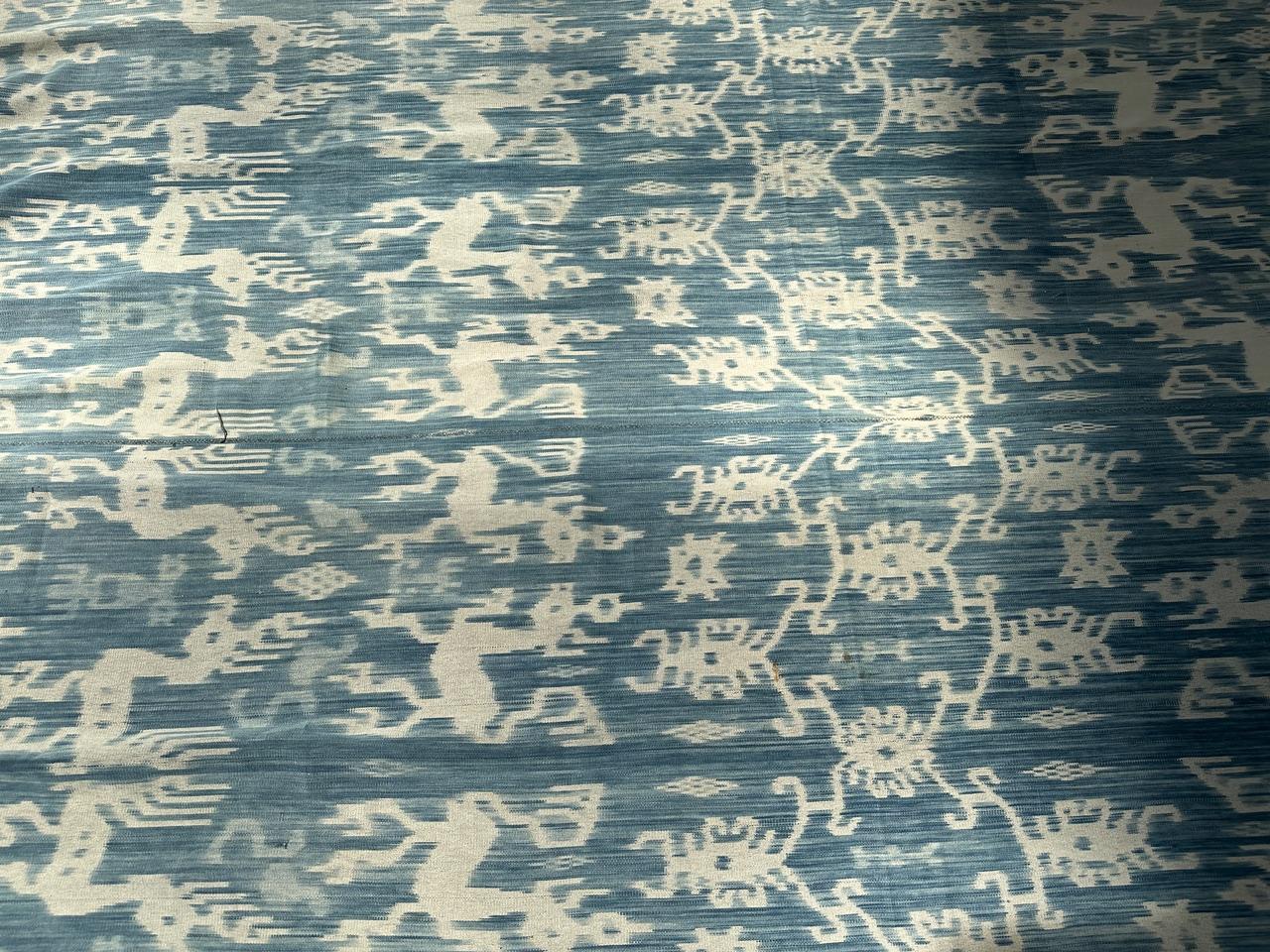 Antico tessuto bianco e indaco proveniente da Sumba. La misura perfetta per essere indossata come uno scialle. Personaggi mitici, cavalli e uccelli si mescolano in questo tessuto grafico. L'ikat è un'antica tecnica utilizzata per aggiungere motivi