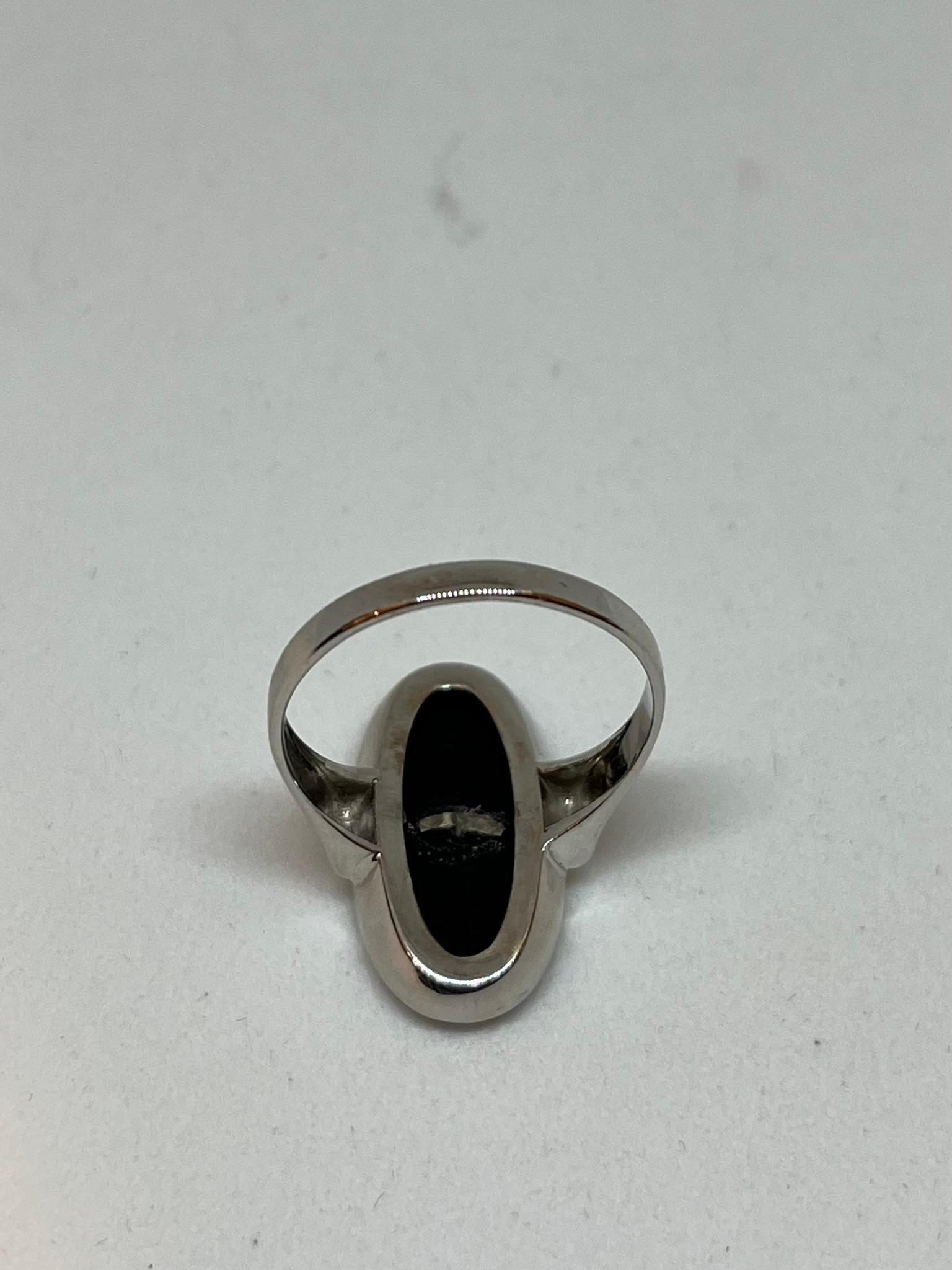 This stunning vintage ring features a classic design, with an onyx stone measuring 20.5 mm x 10.5 mm set in an oval 9 kt white gold setting. A diamond is also set in the center of the onyx, adding a touch of light to the ring.

It presents itself as