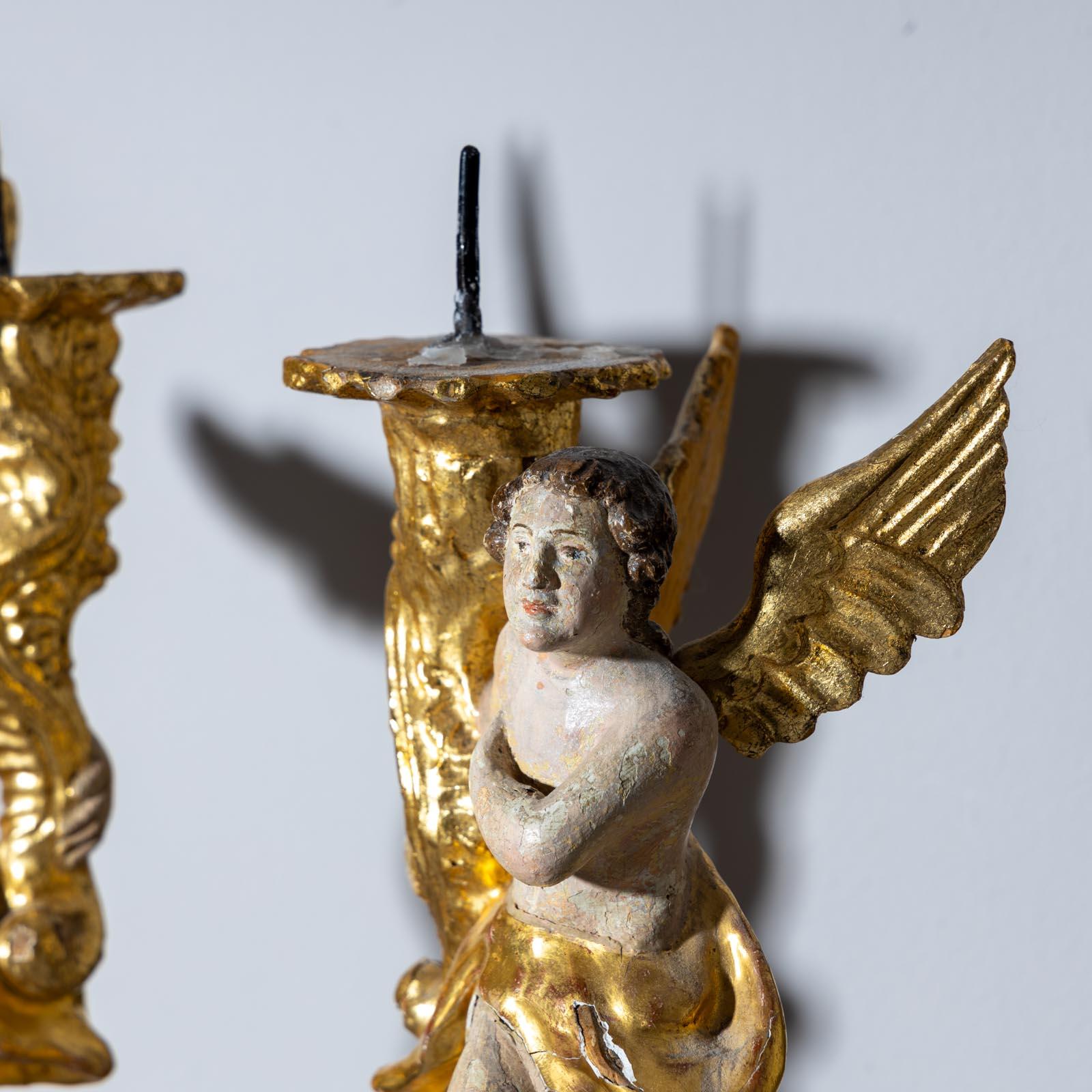 Pair of candlesticks designed in the shape of winged angels. The angels each carry a cornucopia with an iron pin and stand on stylized clouds. The wings, the robe and the candlesticks are gold-patinated. The polychrome setting of the candlesticks is