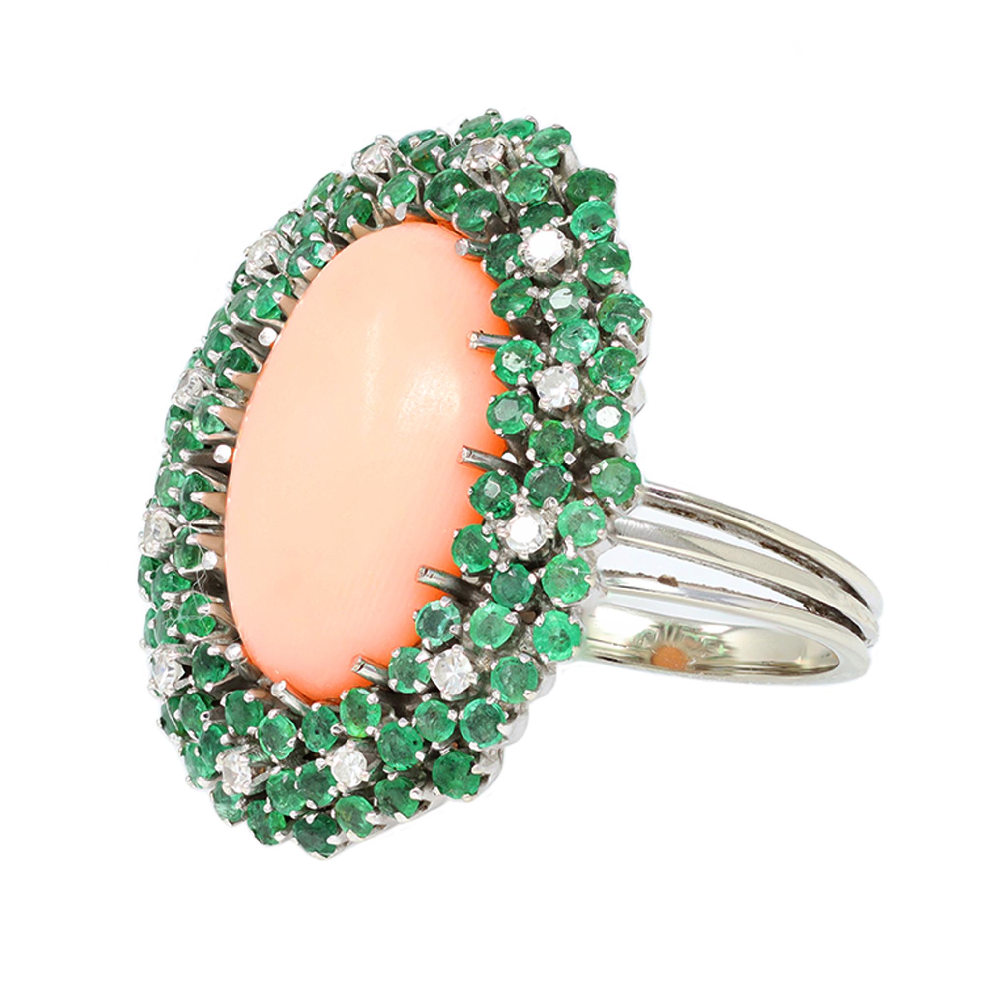 An Italian Angel Skin Coral cabochon ring with emeralds and diamonds set in 18K white gold, circa 1950. Handmade in Italy, this cocktail ring features a blush pink oval shape coral measuring 12x17 millimeter, surrounded by emeralds and diamonds. The