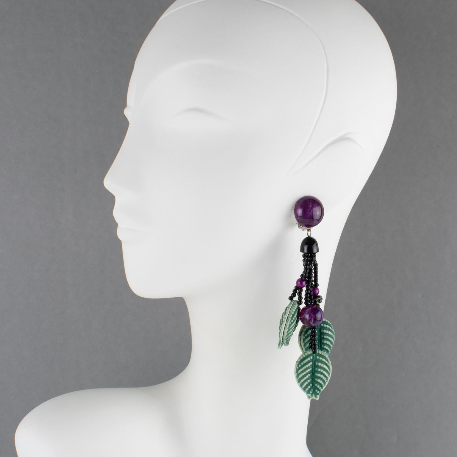 Questi splendidi orecchini a clip in resina di Angela Caputi, made in Italy, presentano una forma lunga, a cascata e penzolante con foglie intagliate e strutturate in modo dimensionale in un colore verde slavato. Sono completati da perline viola