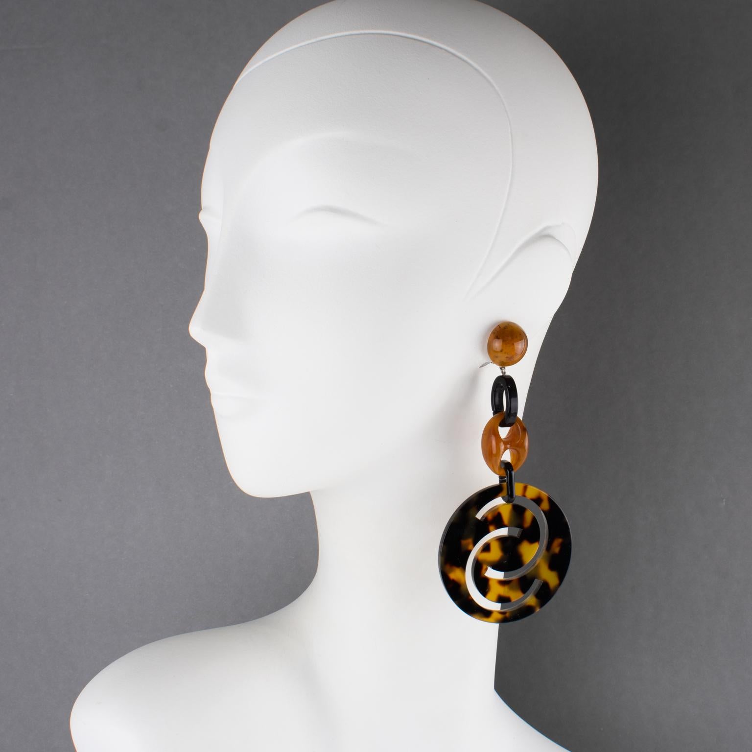 Questi sofisticati orecchini a clip in resina made-in-Italy di Angela Caputi sono caratterizzati da una forma a spalla penzolante con maglie geometriche in resina nera e ambra contrastate da importanti dischi rotondi a spirale intagliati e