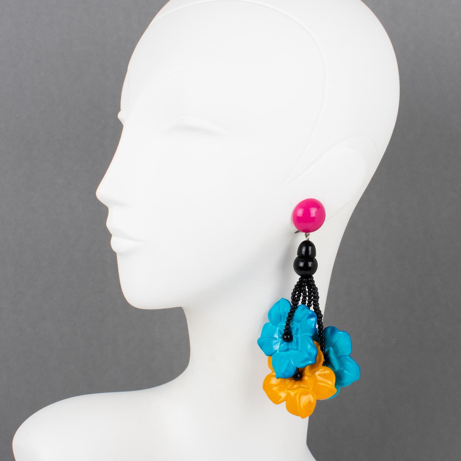 These stunning Angela Caputi, made-in-Italy resin clip-on earrings boast an oversized dangling-drop design with floral elements. The earrings feature three large, intricately carved flower petals in turquoise blue and yellow saffron colors. They are