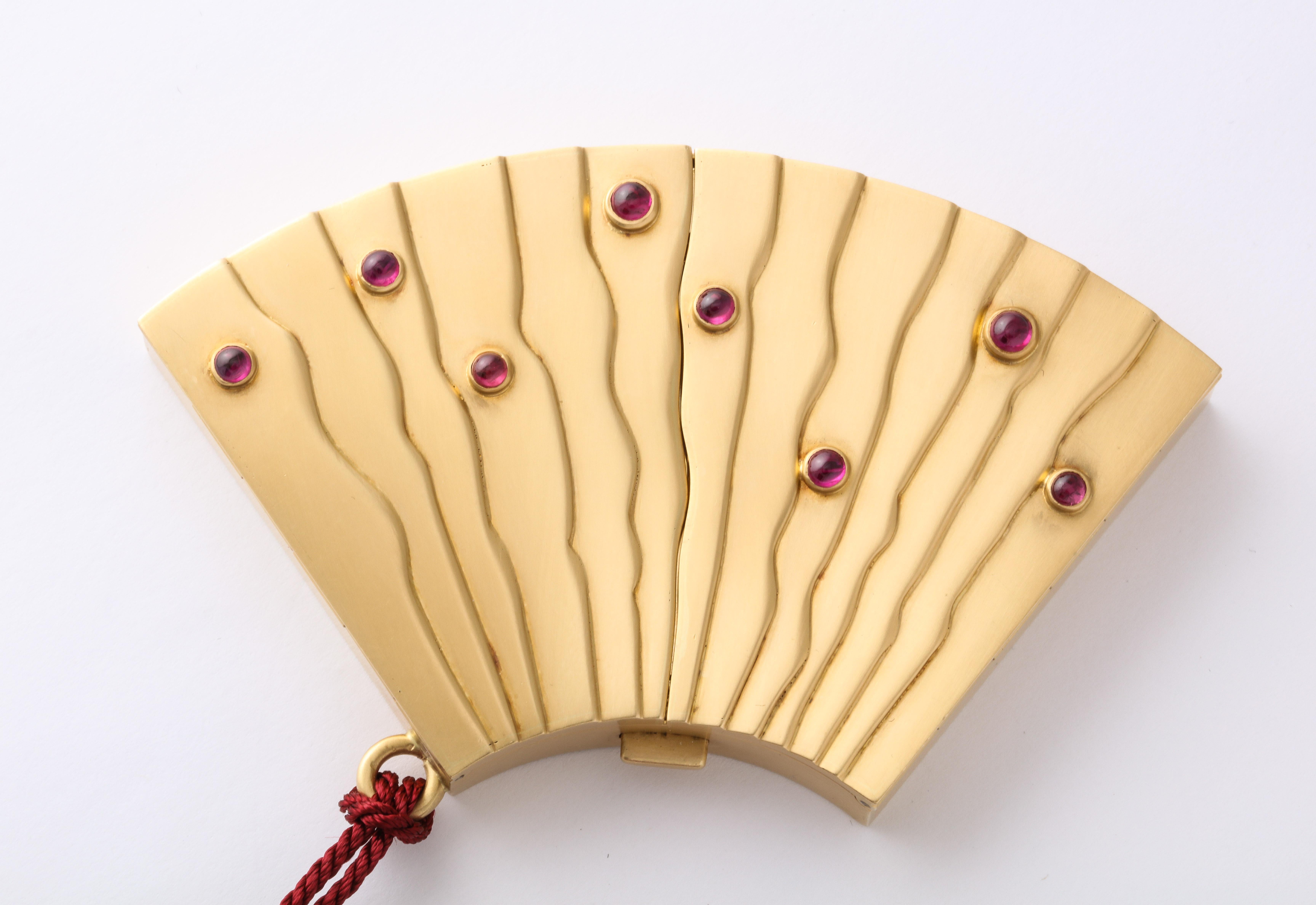 Angela Cummings fan-shaped compact in 18k yellow gold with red tassel, bezel set with 8 cabochon rubies totaling approximately 1 carat. Marked 750, Cummings.  Circa 1970, never used.

Material:
18k Yellow Gold

Stones:
8 cabochon rubies, totaling