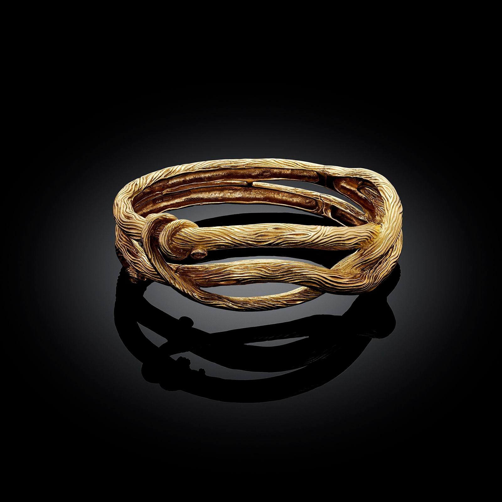 A vintage yellow gold bangle bracelet by Angela Cummings, 1985. The bangle is designed as an organic, twisting style with the gold engraved with fluid lines stylised wood grain. The bangle opens with a side hinge and a concealed tongue and box