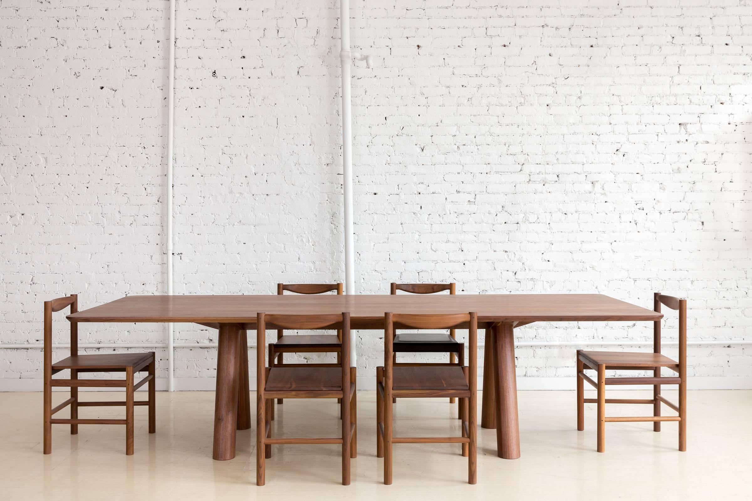 This contemporary, minimal walnut dining table produced in 2017 features large diameter legs and a bold trestle to give rise to an elegant hardwood top. Top includes rounded corners with an optional underside bevel to compliment details found on the
