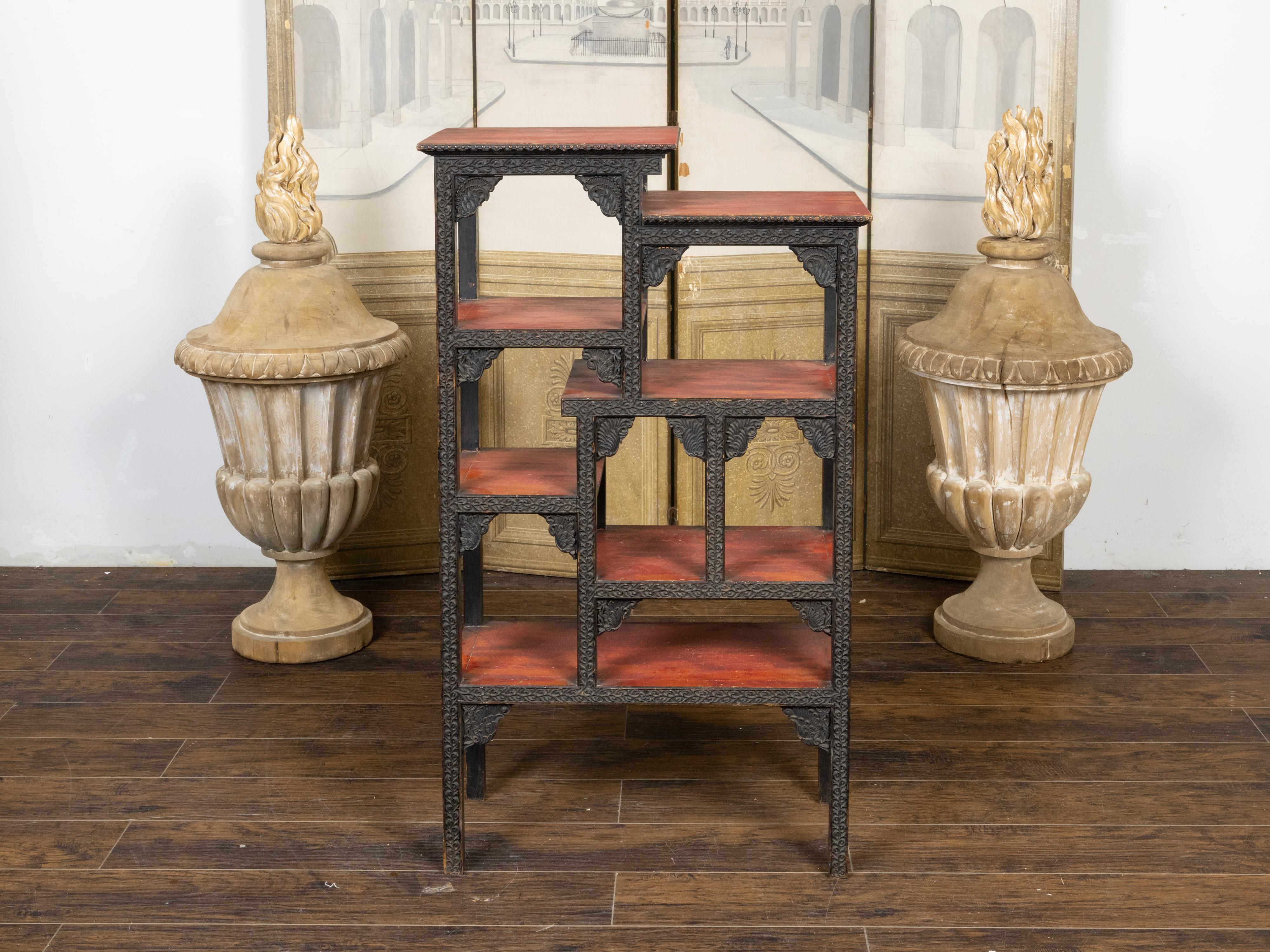 An Anglo-Indian tiered shelf from the early 20th century, with richly carved structure, scrolling foliage, reddish brown accents and black paint. Hand-crafted during the second quarter of the 20th century, this tiered pieces features seven shelves