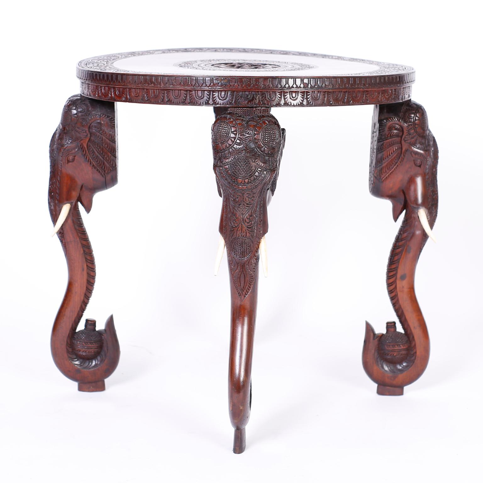 Anglo Indian table or stand crafted in mahogany with a round top having a carved center medallion depicting a deity with elephants surrounded by floral carvings. The four elephant head legs have bone tusks and trunks carrying flasks.