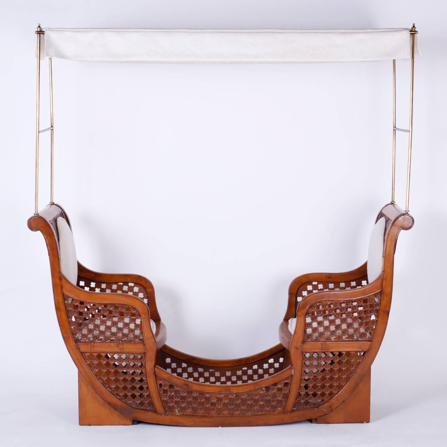 Antique elephant seat or howdah converted to a tête à tête. Featuring a brass canopy frame, upholstered seats at each end of a graceful mahogany frame carved with flowers, and a floral carved trellis on the sides. If only furniture could talk, what