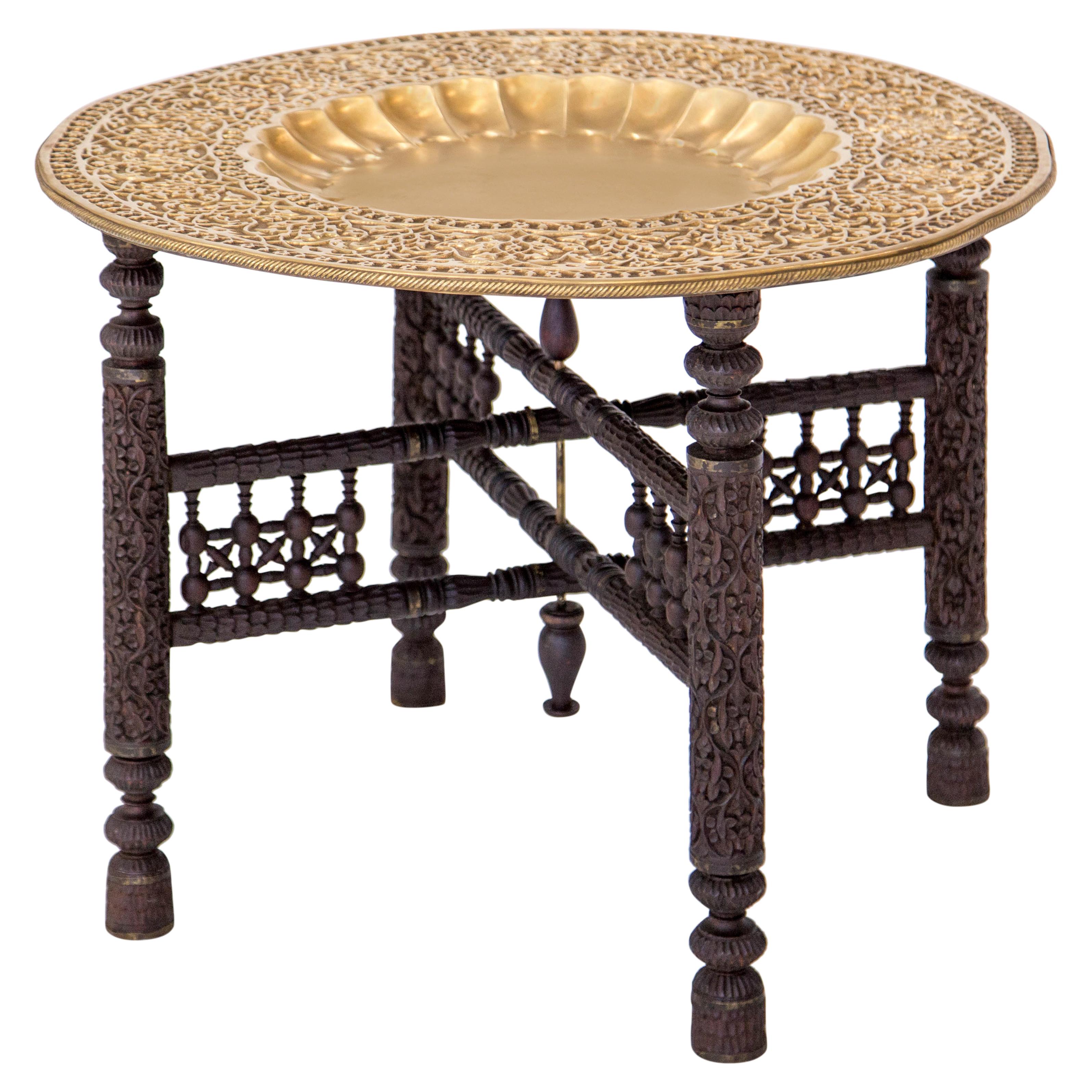 Early 20th century solid brass tray coffee on a folding carved wood stand. The tray has a smooth dished center with scalloped sides surrounded by an wide ornate stamped rim in a geometric foliate pattern inside a subtle pie crust edge. The tray