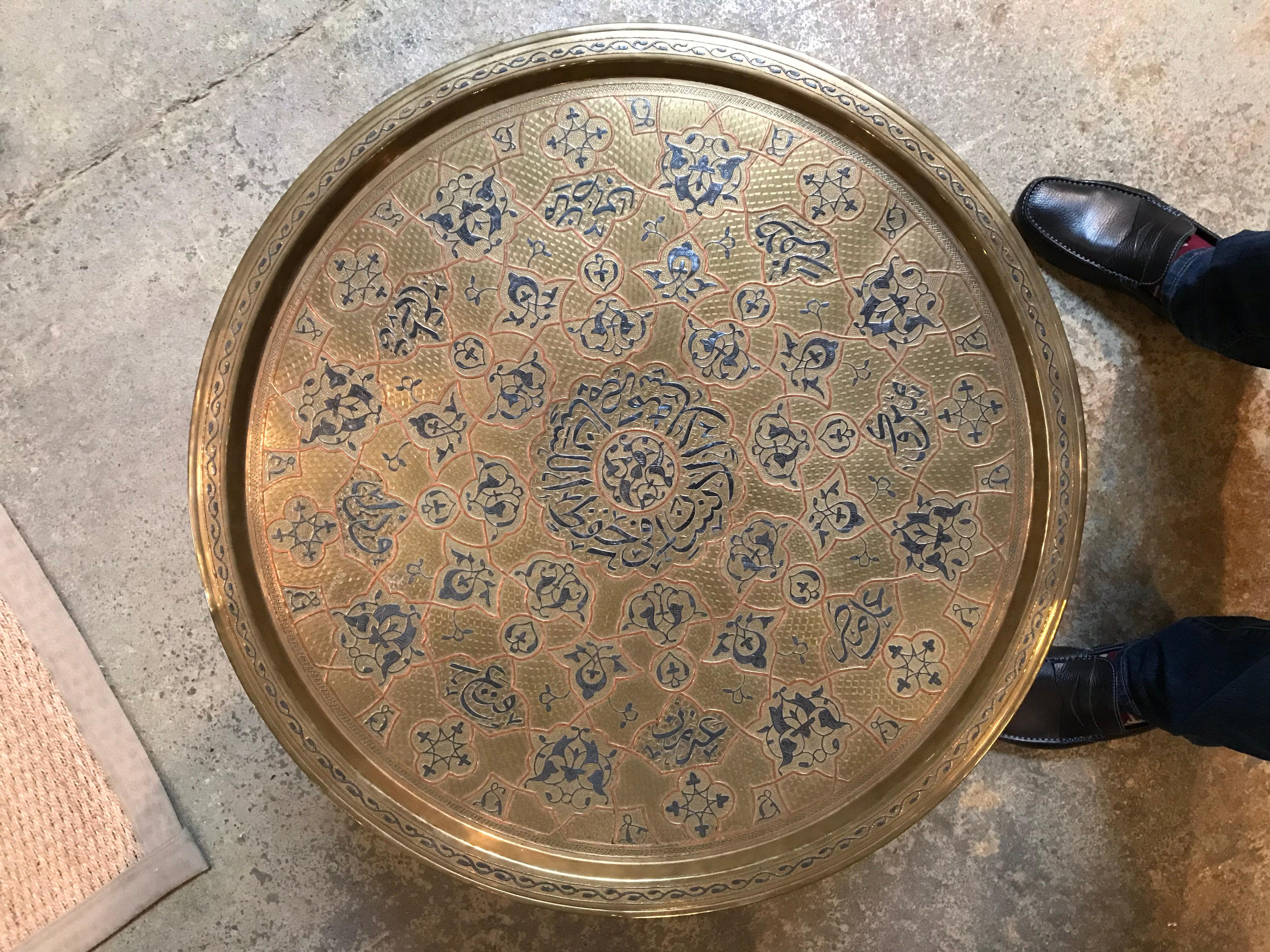 This Anglo-Indian table has a folding base that goes from 6 legs into 2. It was made in the 19th century and includes a round metal tray as the tabletop. The tray has blue patterns with red outlines.