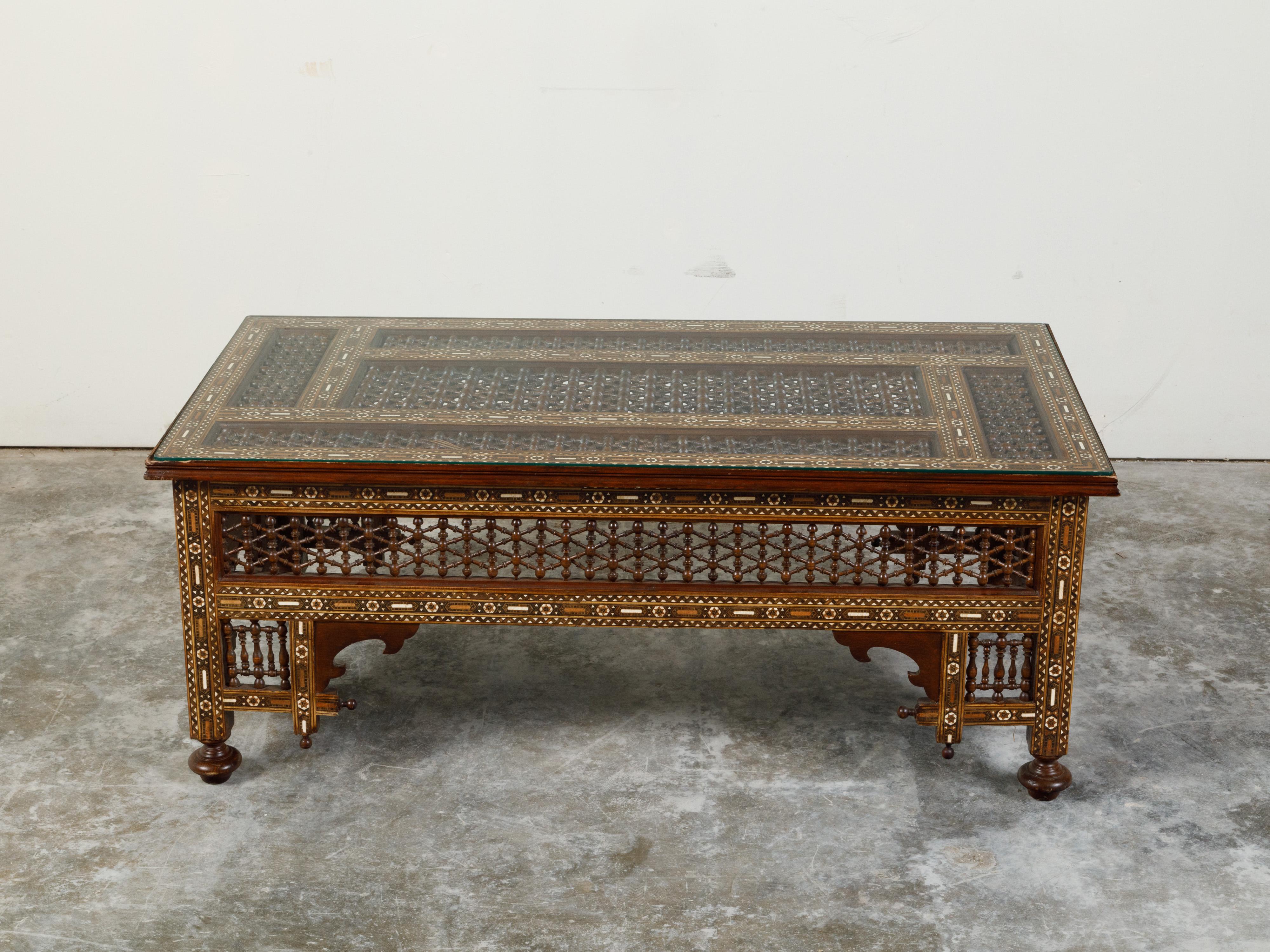 An Anglo-Indian glass top coffee table from the early 20th century, with mother-of-pearl geometric inlay and intricate carved motifs. Created in India during the first quarter of the 20th century, this coffee table features a rectangular glass top