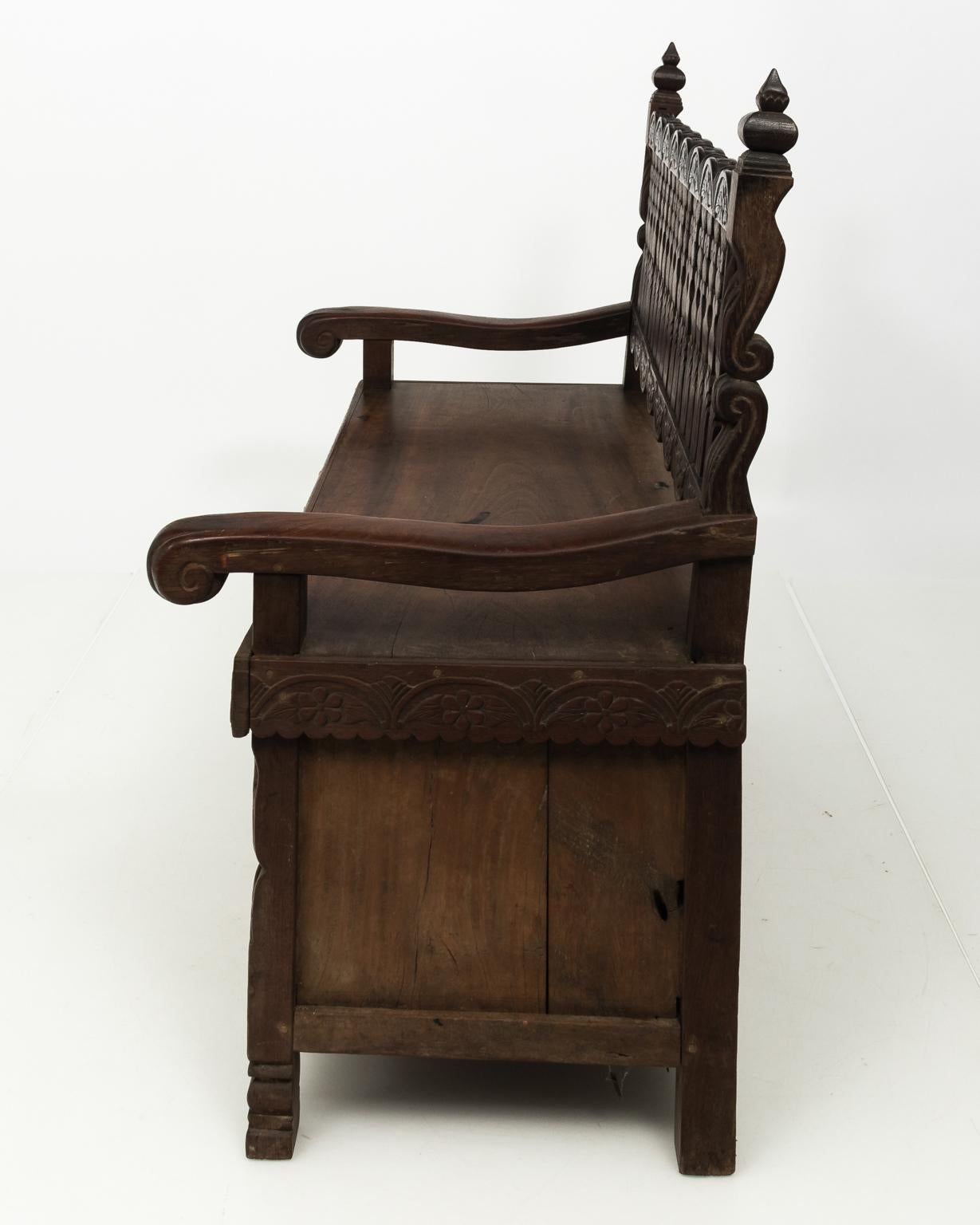 Anglo-Indian hall bench in teakwood with carved back splats and storage below the seat behind panels. The bench also features carved floral trim and scrolled arms, circa 19th century.
 