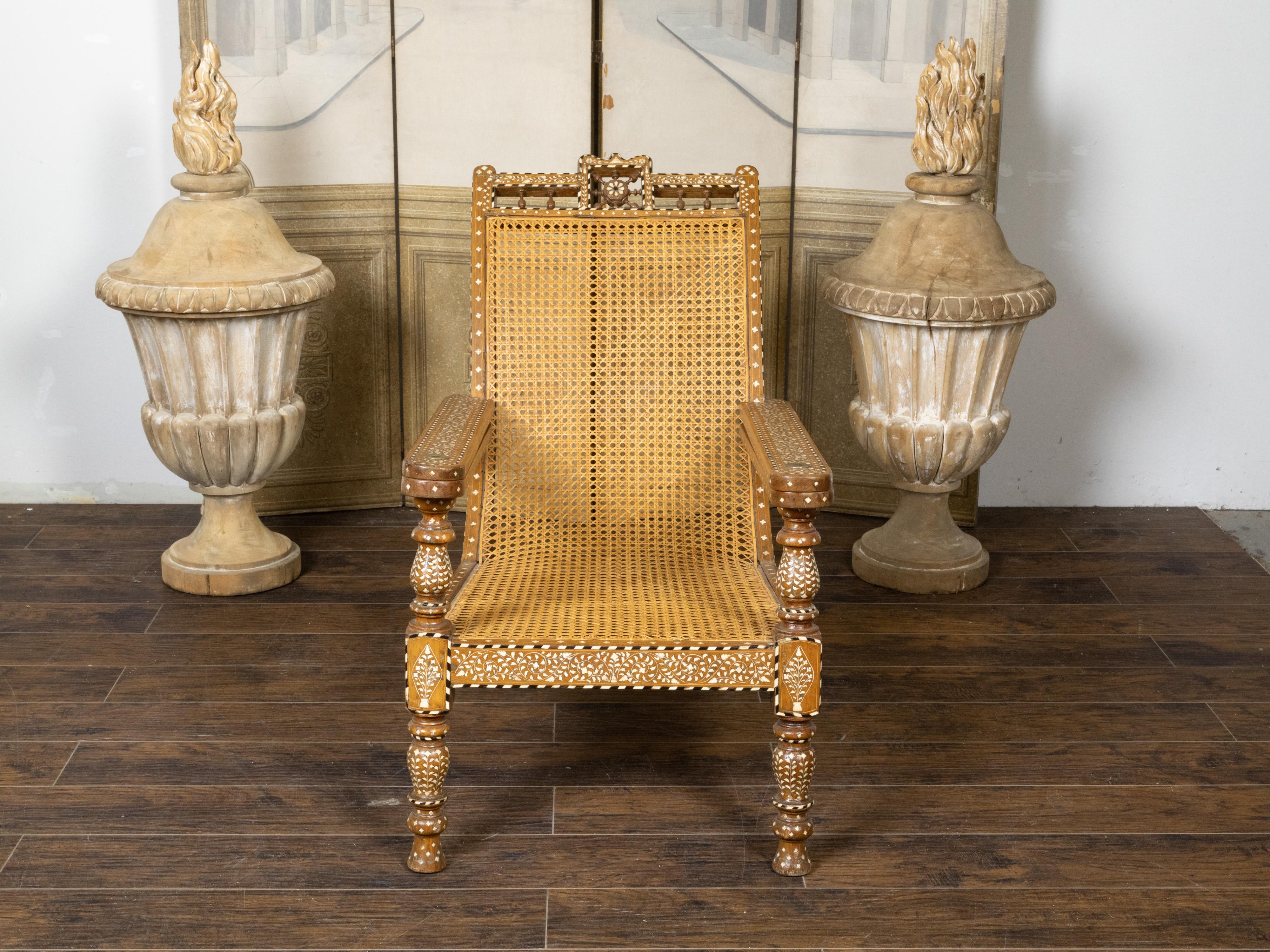 An Anglo-Indian wood and cane plantation chair from the early 20th century, with foliage-themed bone inlay and extending arms. Created during the early years of the 20th century, this Anglo Indian plantation chair features a slanted back topped with