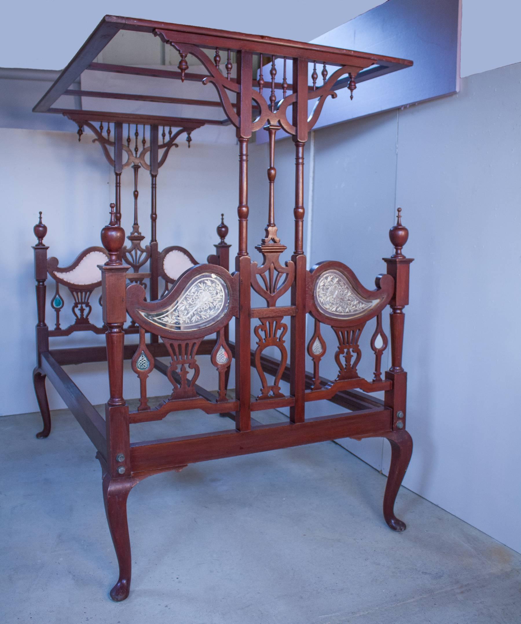 A richly toned mahogany bed with tester canopy from northern India, circa 1930. The intricately carved frame has mirrors reverse etched with musical instruments on the outside of the head- and footboards and upholstery on the inside. The frame comes