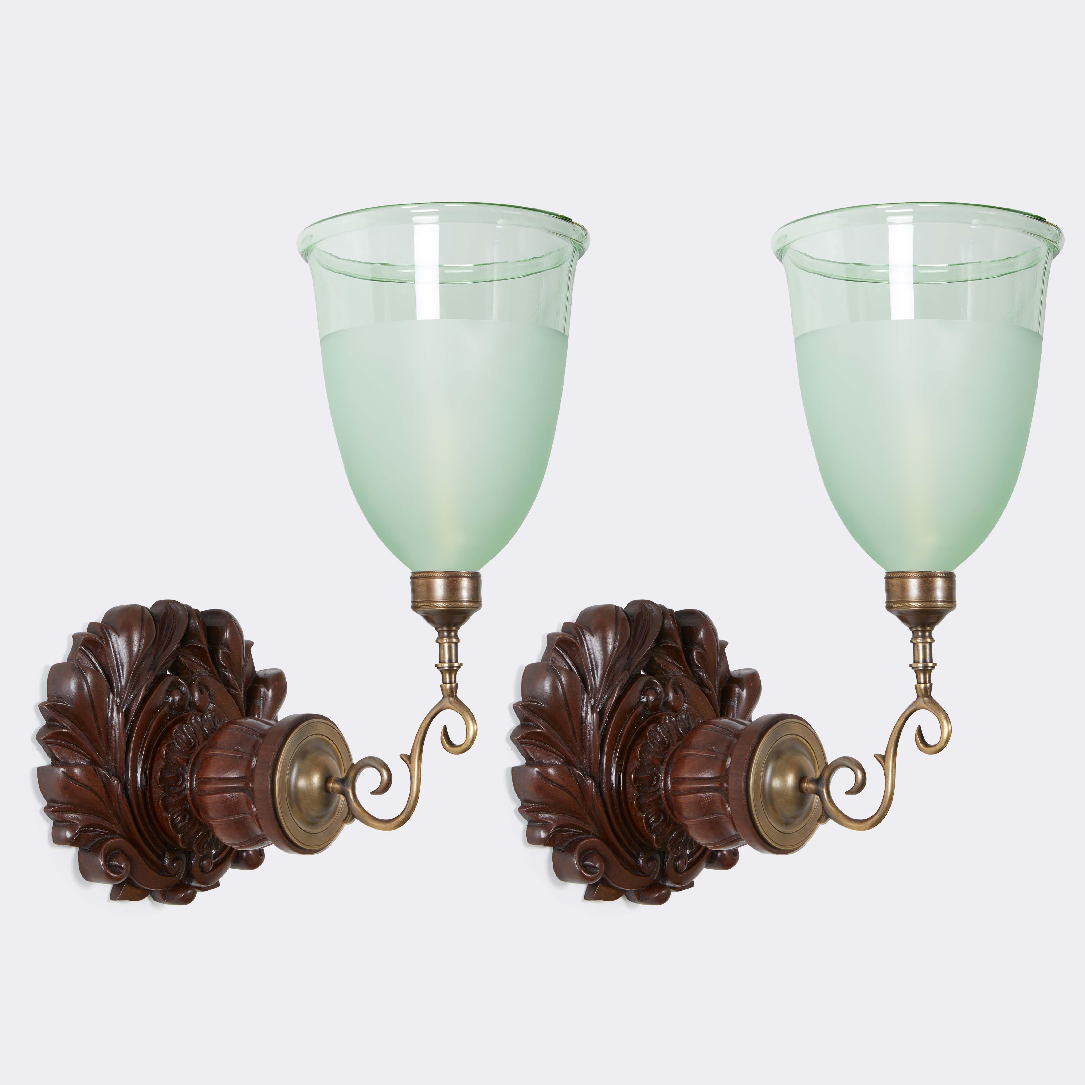 A pair of single light Anglo Indian styled sconces with antique backplates. The hand carved rosewood backplate with acanthus leaves and lotus shaped element issuing the round backplate with candle arms securing hand blown green hurricane shades,