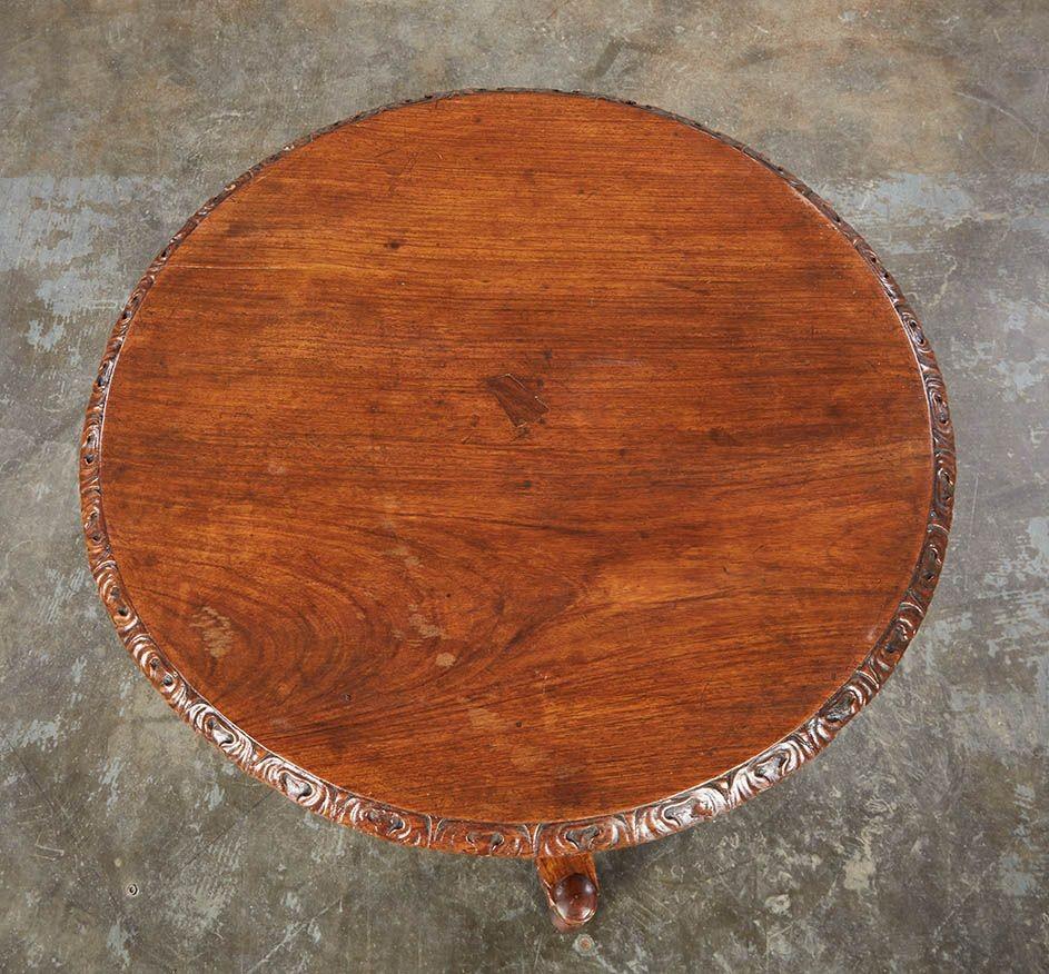 Mesa angloindia de madera de palisandro con tablero redondo y borde y delantal tallados con motivos foliados, sobre pedestal de pétalos de tulipán igualmente tallados con mecanismo basculante, que termina en una base trípode tallada y perfilada