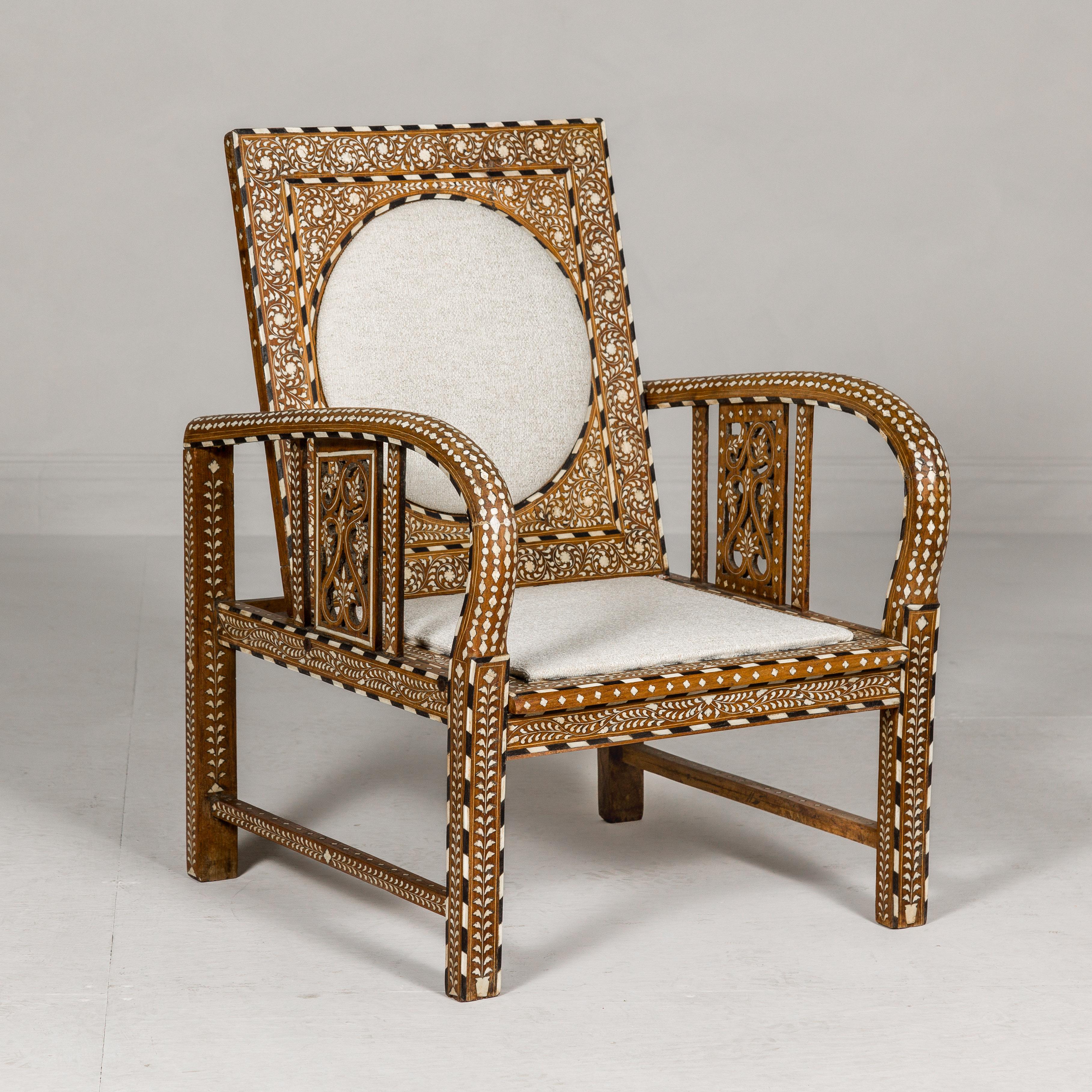 An Anglo-Indian style mango wood armchair with scrolling foliage bone inlay, loop arms and custom upholstery. This Anglo-Indian style mango wood armchair is a true masterpiece, a blend of exquisite craftsmanship and opulent design. Adorned with