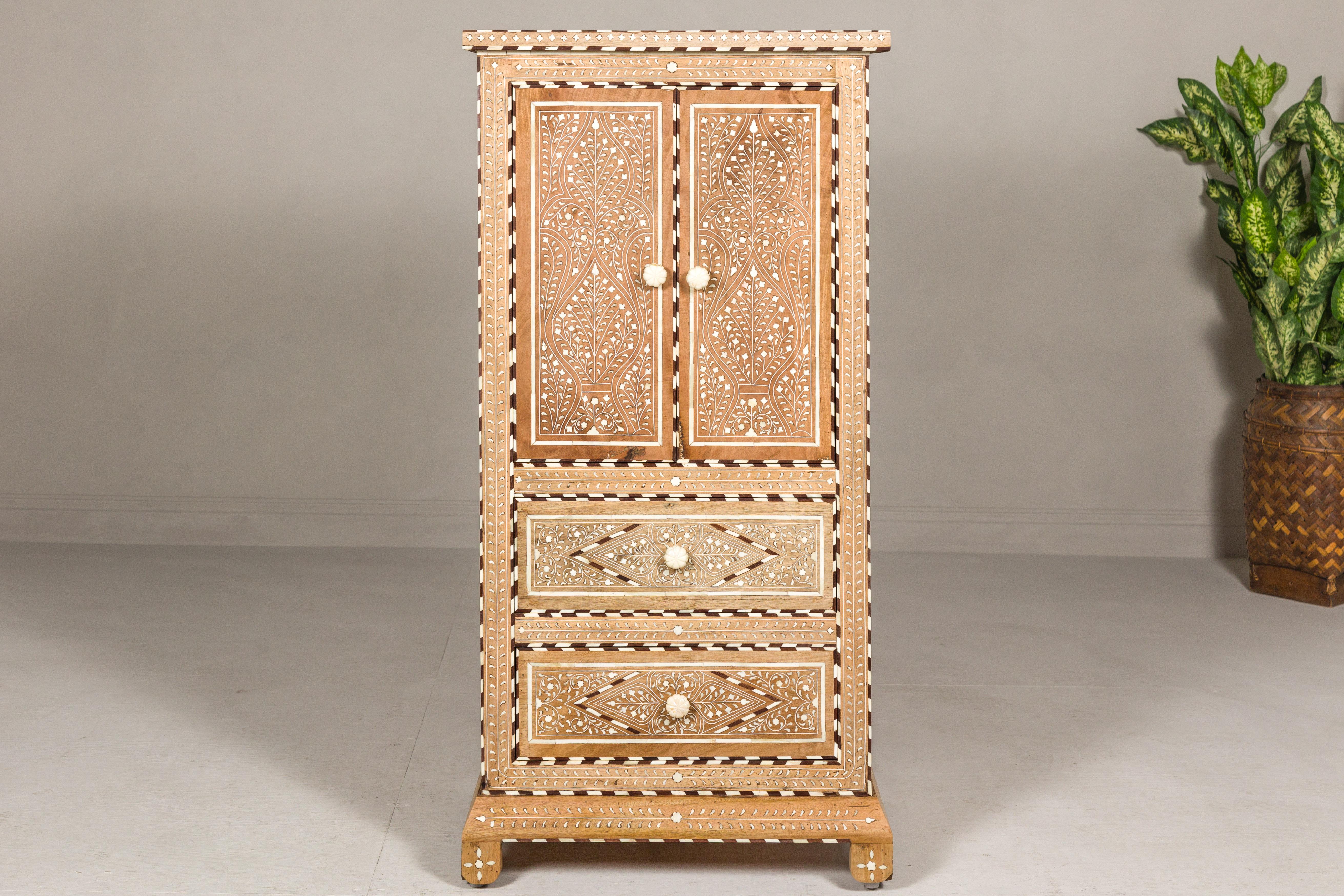 An Anglo-Indian style narrow mango wood cabinet with foliage-themed bone inlaid décor, two doors, two drawers, bone hardware and natural patina. Behold the artisanal mastery of this Anglo-Indian style mango wood cabinet, a true testament to timeless