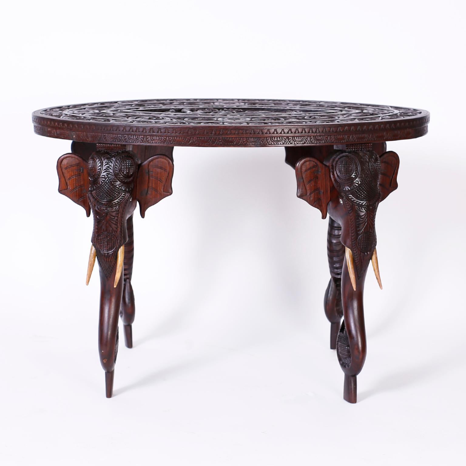 Antique Thai table crafted in indigenous mahogany with an elaborately carved oval top with a goddess in the center panel inside three layers of floral designs. The four legs are carved elephant heads with wood tusks and stylized feet.