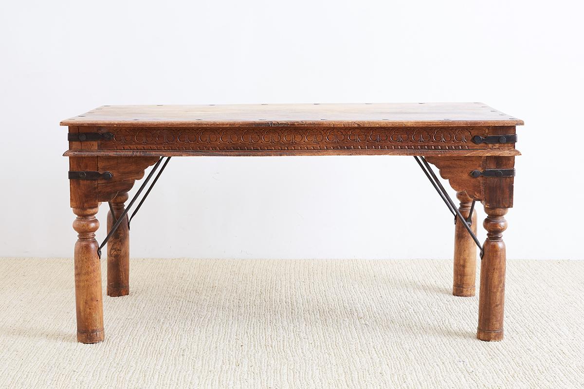 Rustic Anglo-Indian dining table or library table constructed from teak. Carved from solid wood with handmade iron nails and iron strapping. The plank top has nails around the border and decorative geometric carvings on the frieze. The table is