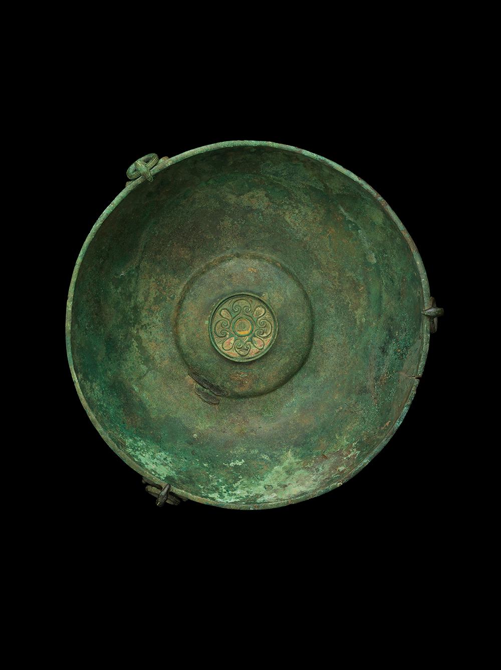A very rare and near-complete copper-alloy hanging bowl and associated fittings. Crafted from a single sheet of bronze, the body of the bowl is curved, with a slightly recessed lip. The bowl features three matching hooked escutcheons and suspension