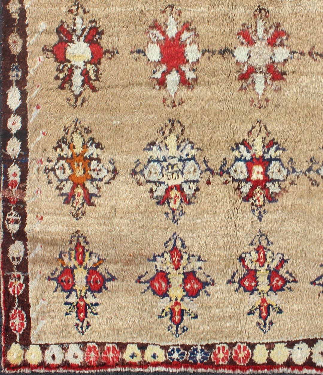 This Tulu carpet features several multicolored floral motifs laid across a sand-colored field and enclosed within a chocolate brown border of alternating botanical motifs. Accent colors include light green, orange, ivory, red and charcoal. Tulu