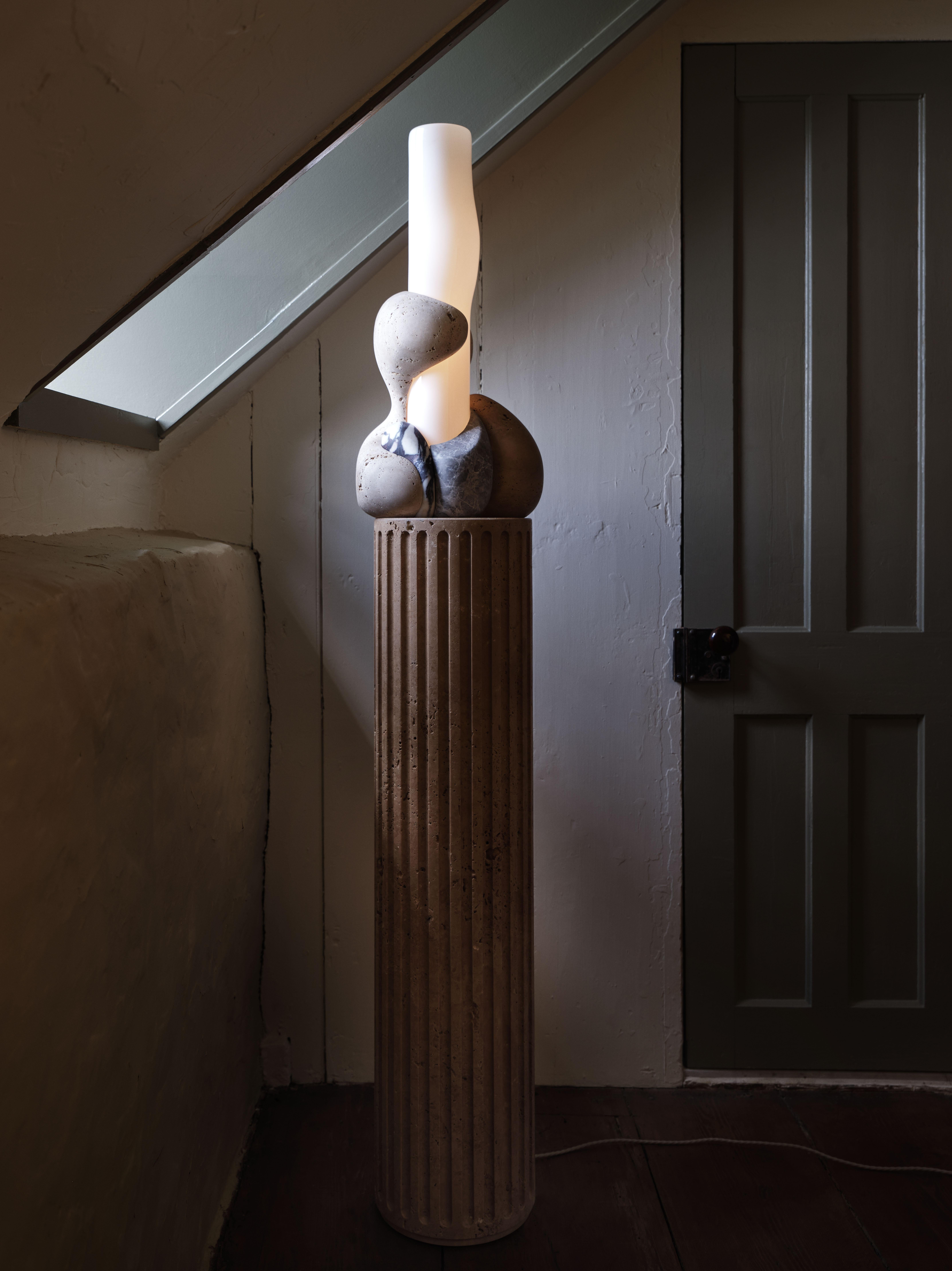 Perched atop of a round fluted travertine plinth, organic forms carved from various types of stone embrace a hand-blown glass cylinder which glows from within.

Materials: Light and Dark Travertine, Siylon Gray, Viola Calacatta, White Glossy