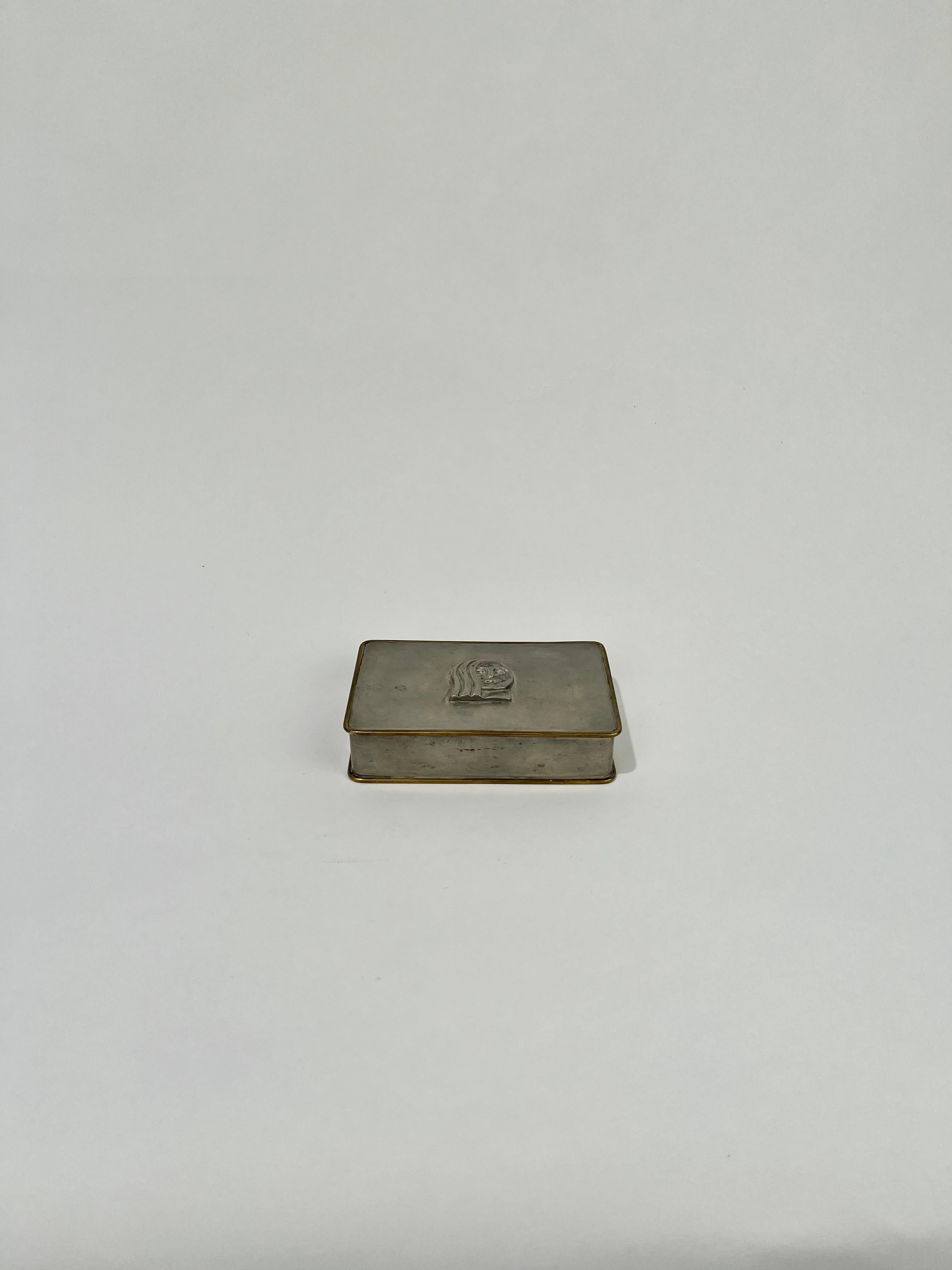 Box in brass and pewter with decor of a womans face. 
Designed by Anna Petrus for Firma Svenskt Tenn. Marked B8 = 1928.

Anna Petrus was a sculptor and designer at the beginning of the 20th Century. After her death, Anna Petrus' work was almost