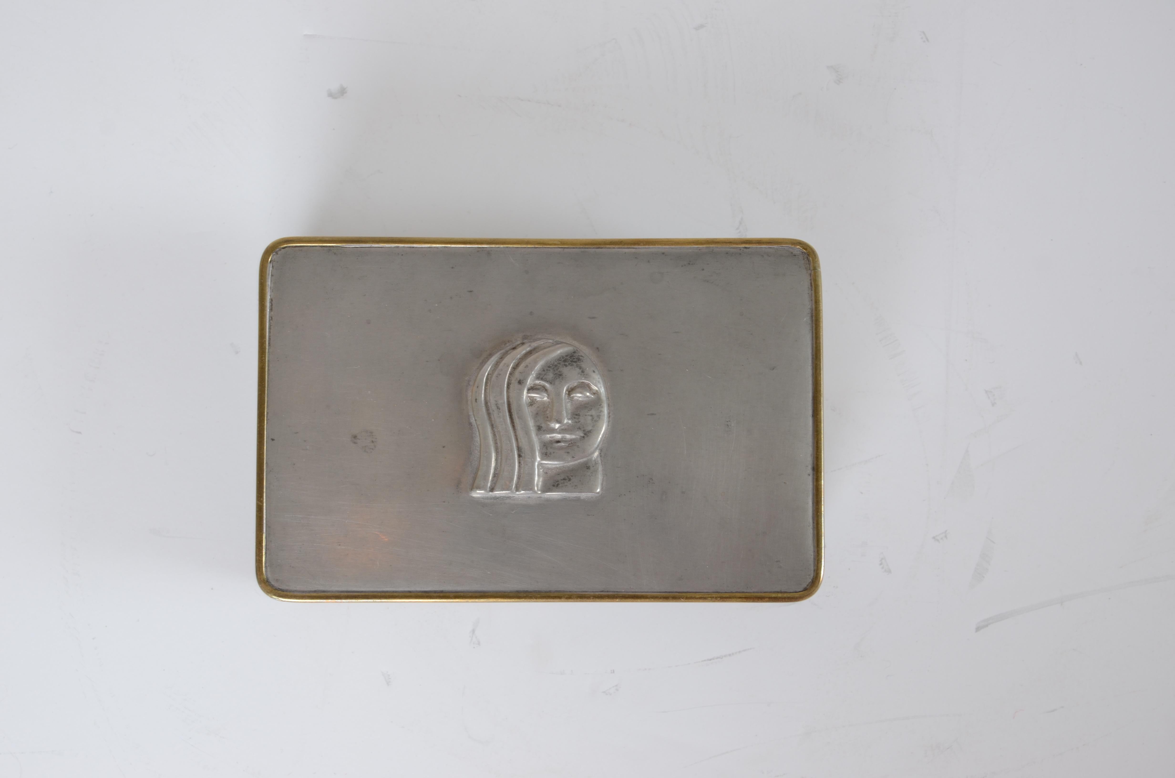 Dose aus Messing und Zinn mit dem Dekor eines Frauengesichts. Entworfen von Anna Petrus für die Firma Svenskt Tenn. Kennzeichnung B8 = 1928.
  