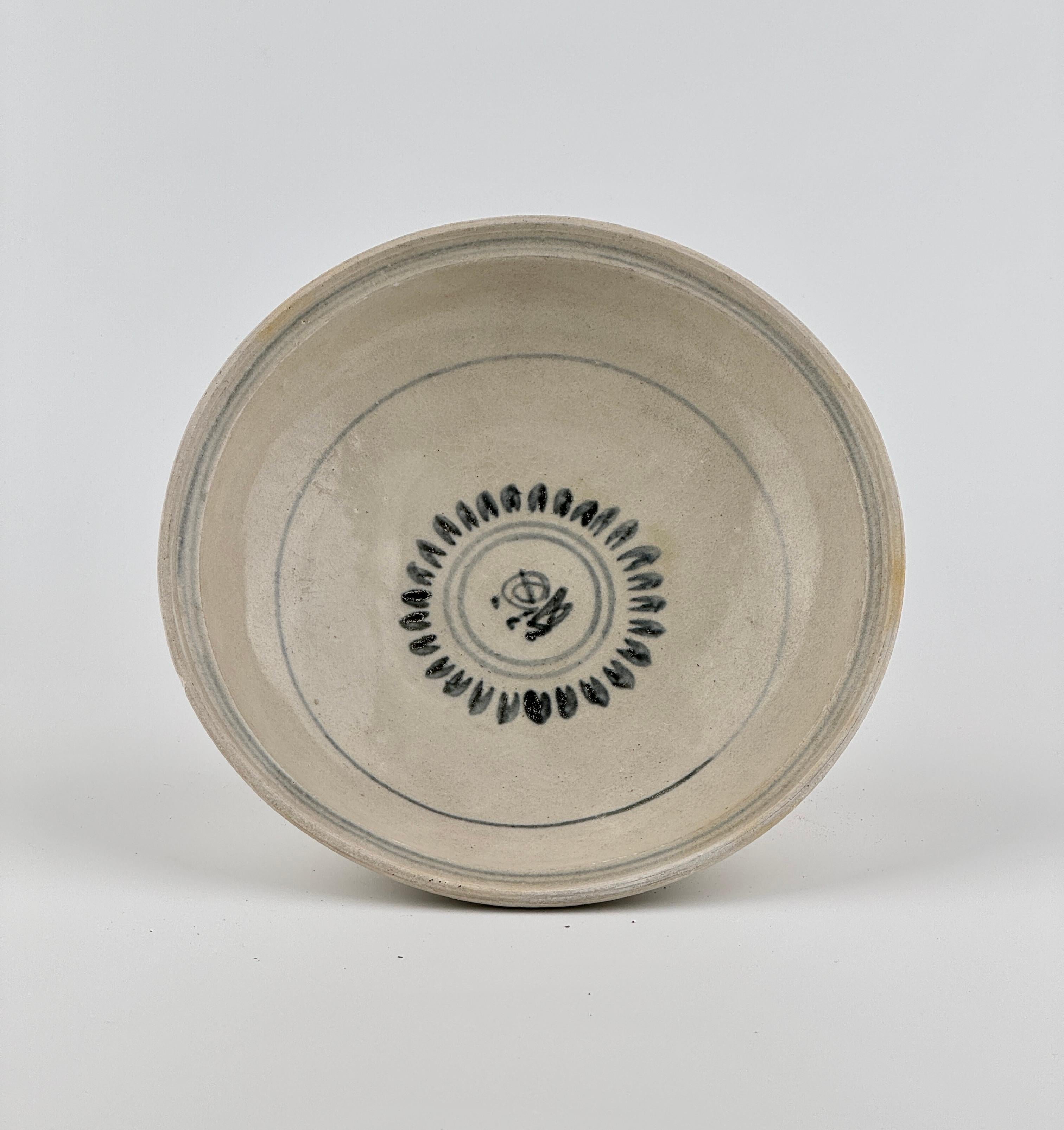 Its surface is coated with a smooth, glossy white or cream-colored glaze, exuding a clean and elegant finish. At the center, a geometric motif painted in black pigment draws the eye, surrounded by a repetitive pattern of leaf-like or feather-shaped