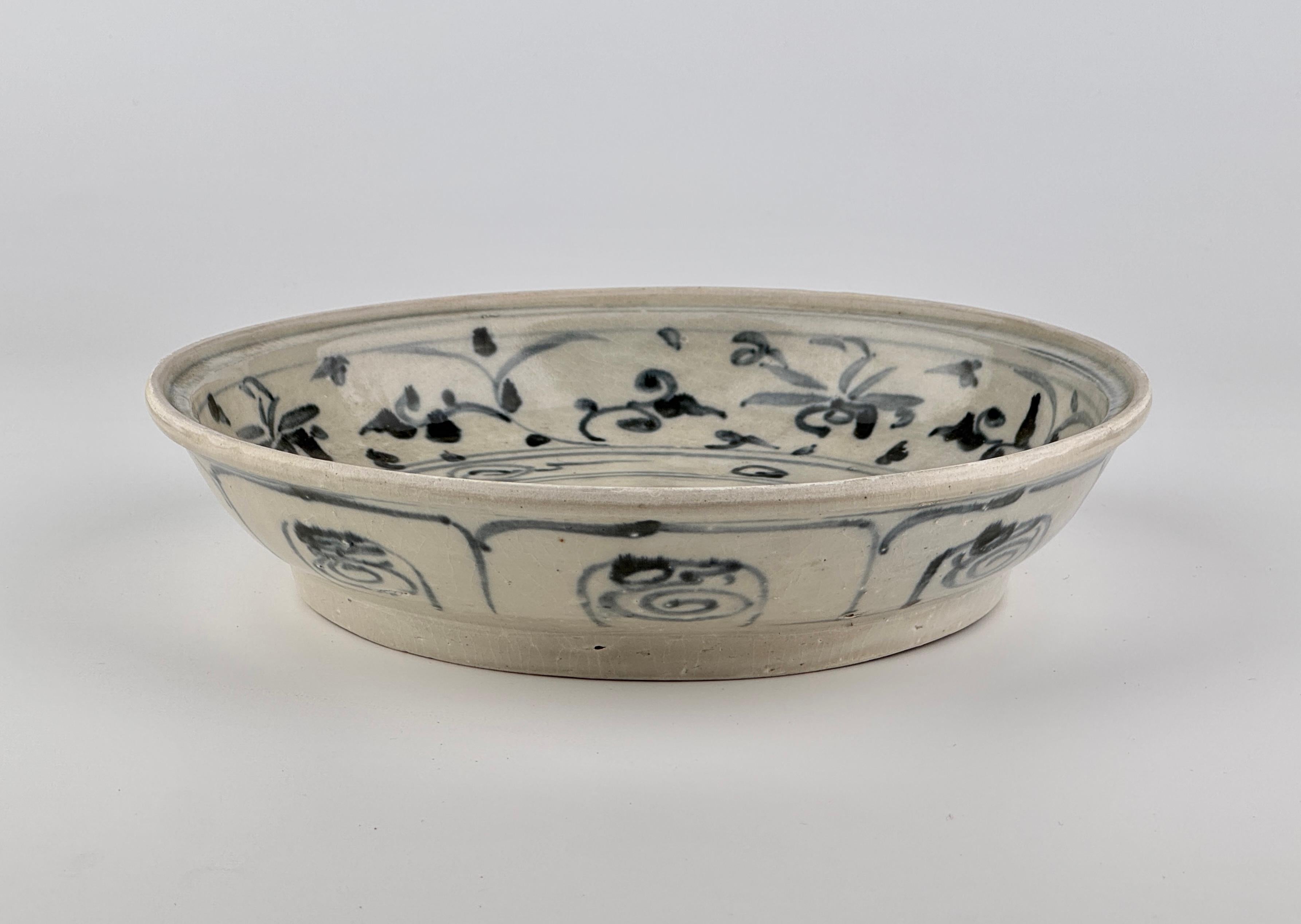 This Annamese ceramic dish features a central lotus motif, enclosed by a thin circular border. The rim is adorned with intertwining vine patterns and swirling motifs.  The dish has a gently curved form with a well-proportioned depth and a neatly