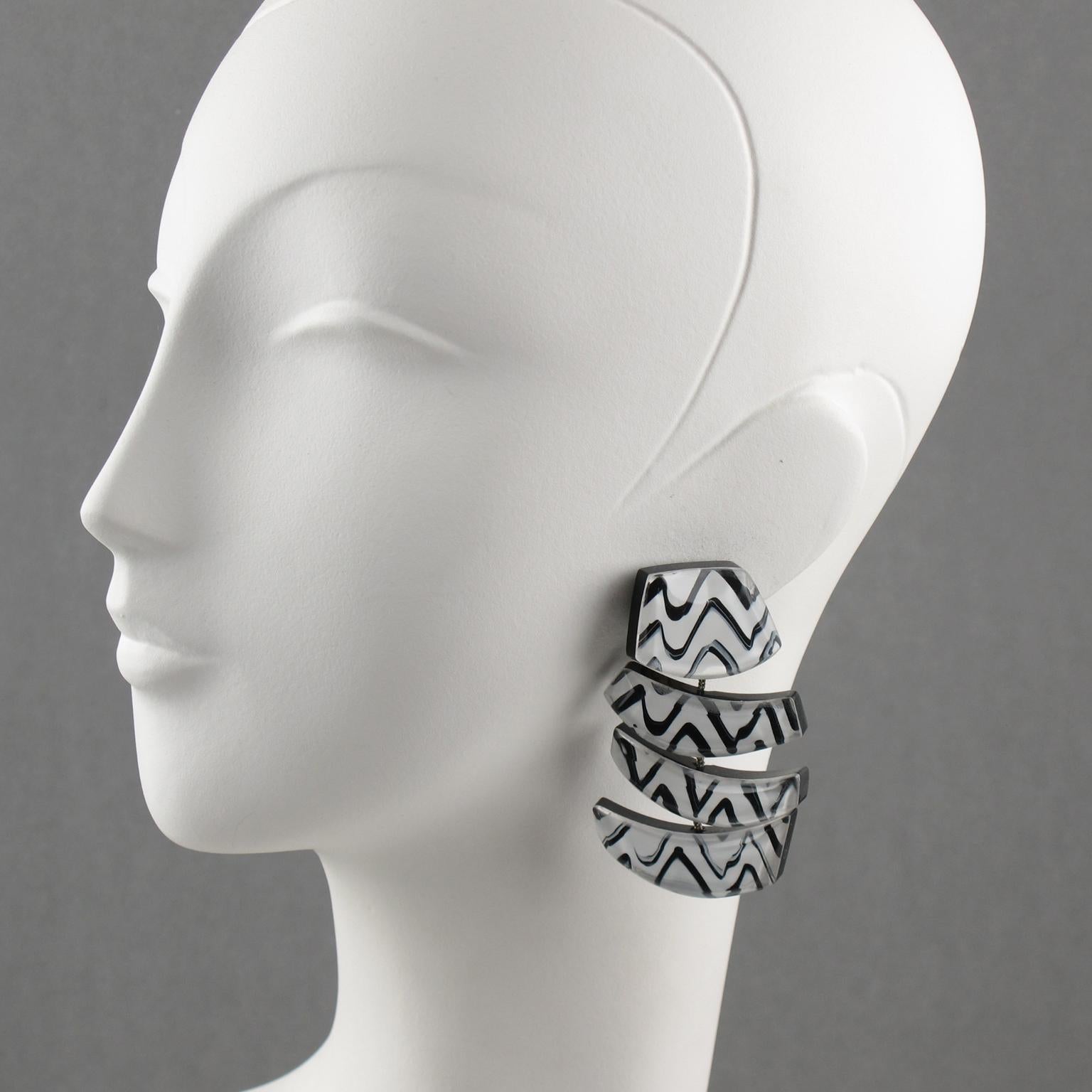 Stunning oversized Lucite clip-on earrings designed by Anne & Frank Vigneri. Huge geometric dangling shape featuring dimensional layer with black and white zig-zag design inclusions and large beveling. The elements are all linked with silver-tone