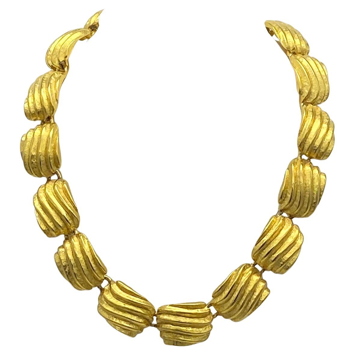 Questa è una collana a maglie in oro di Anne Klein del 1990. È composto da quadrati convessi variegati e strutturati in tonalità oro brillante. Questa collana regolabile in lunghezza è dotata di tre anelli e un alamaro, oltre a un charme con il logo
