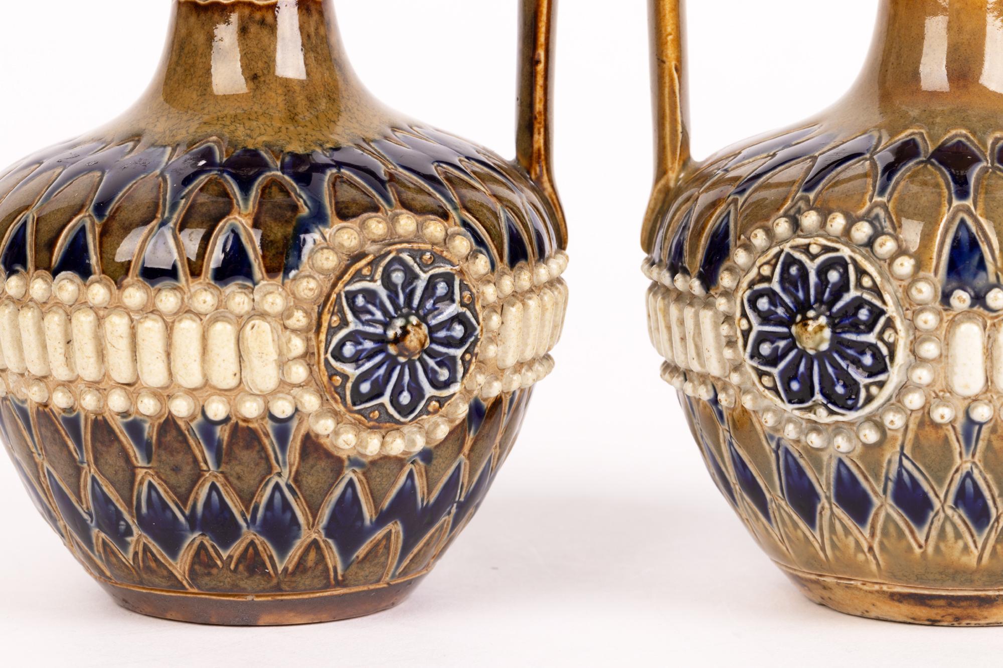 A good and decorative pair antique Doulton Lambeth stoneware jugs decorated with stylized floral designs by Annie Cupit and Florence Dennis and dating from around 1891. The jugs stand on a flat unglazed base with squat bulbous rounded bodies, a