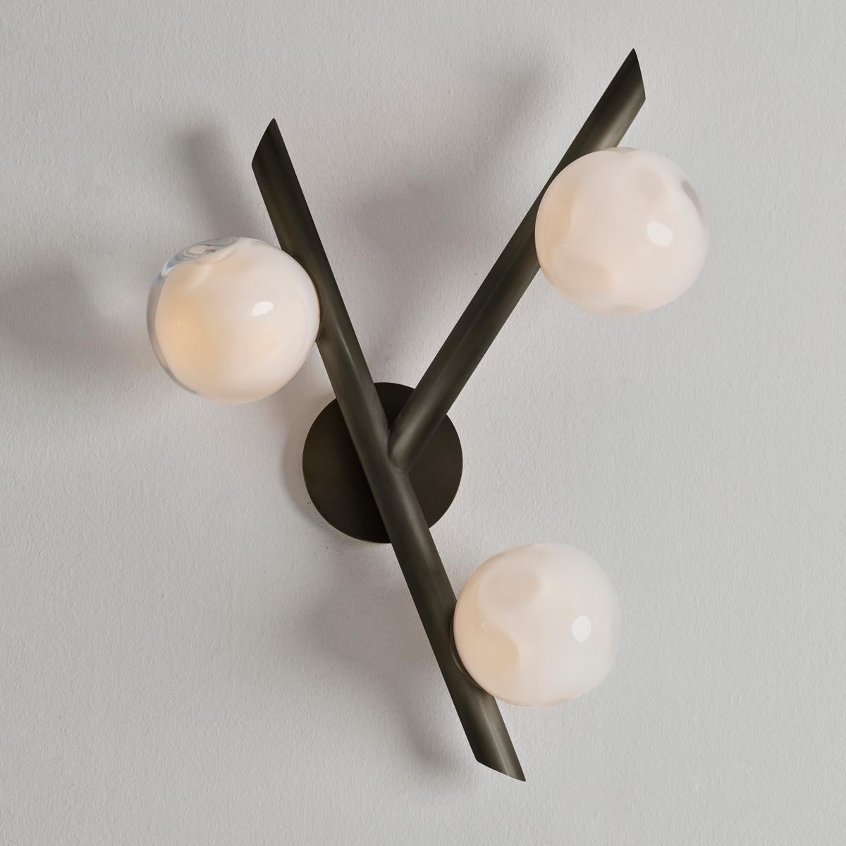 The Antares wall light harmoniously balances the linear details of its intersecting arms with the organic form of its handblown glass shades. The first images show the fixture in our Brunito Nero (Black bronze) finish-subsequent pictures show it in