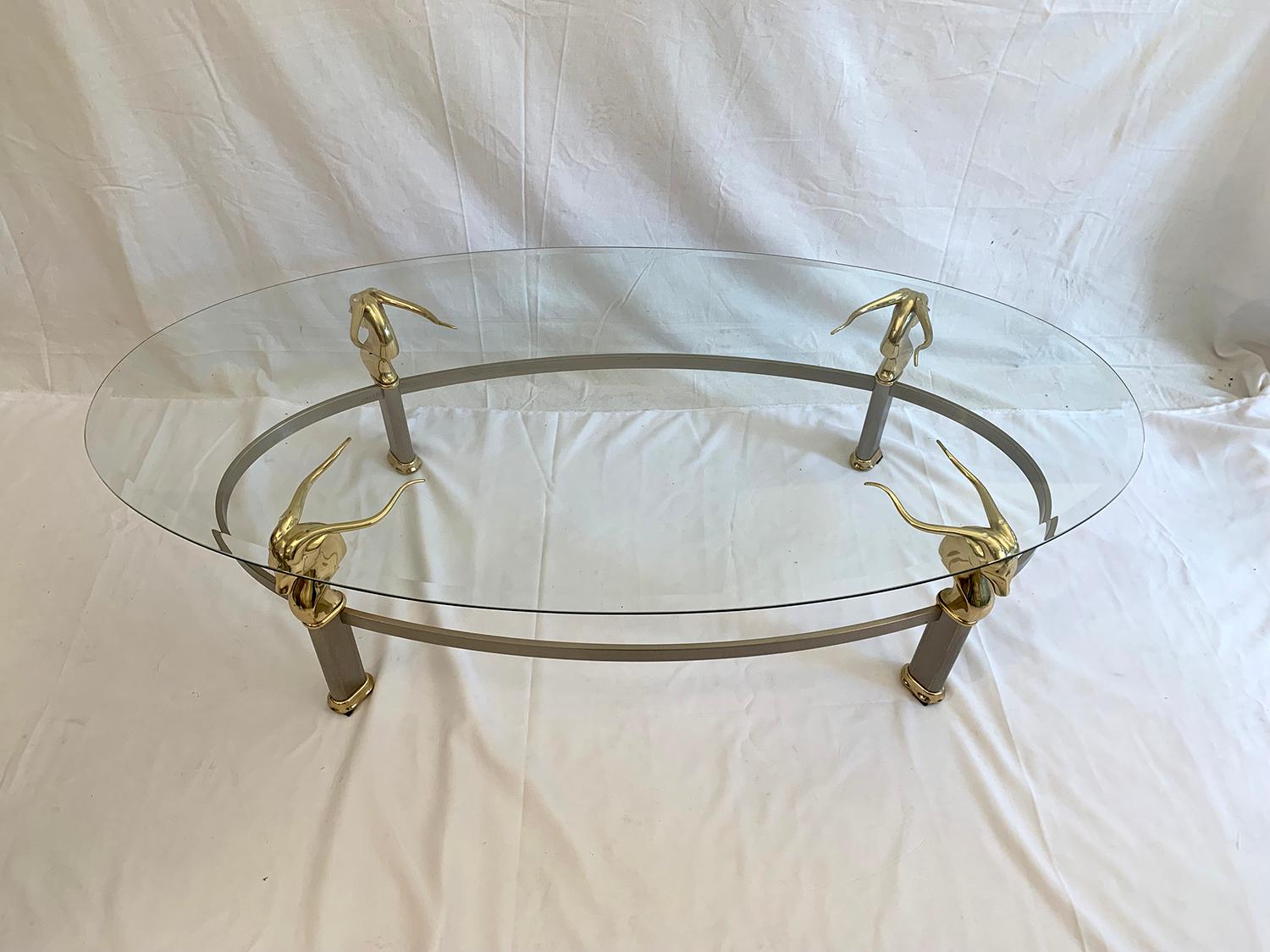 Very beautiful and rare coffee table made of an oval structure in brushed metal, adorned with polished gilt metal antelope heads and with a transparent glass on its top. This table is in very good general condition for his age, there are just some